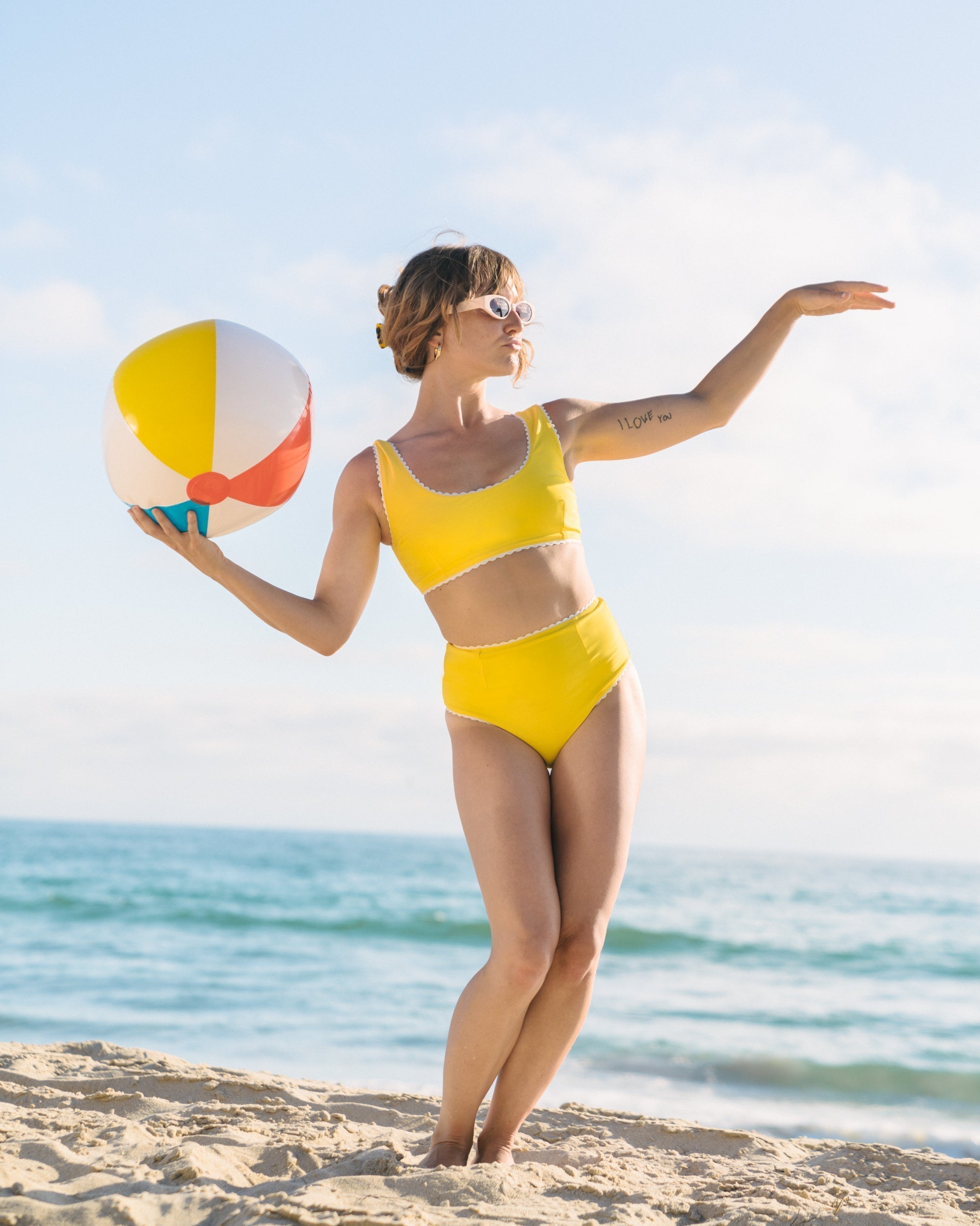 Woman in a yellow bikini holding a beach ball on a sandy beach with ocean and sky in the background