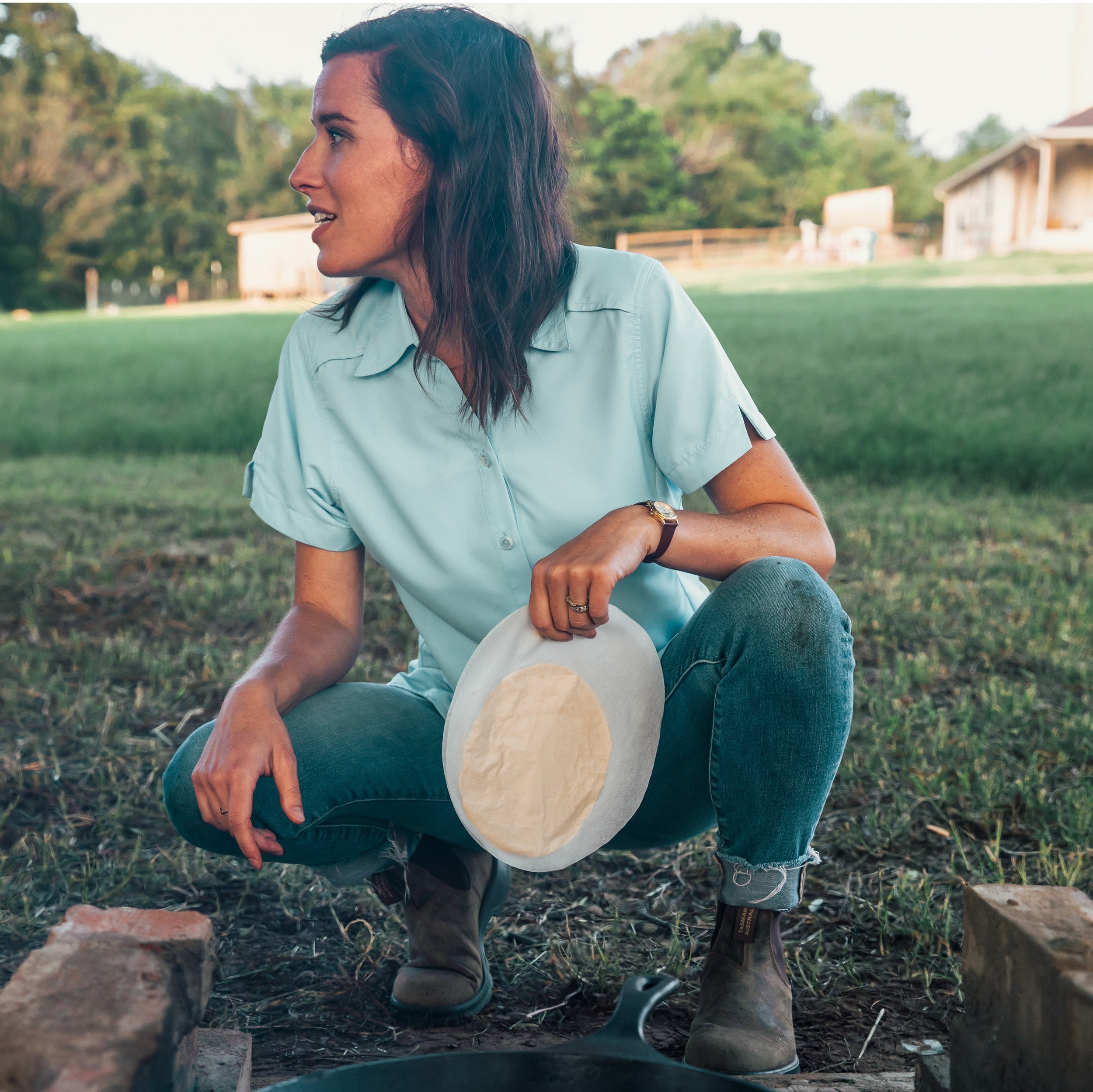 Woman in a light blue shirt and jeans holding a white disc in a grassy outdoor setting.