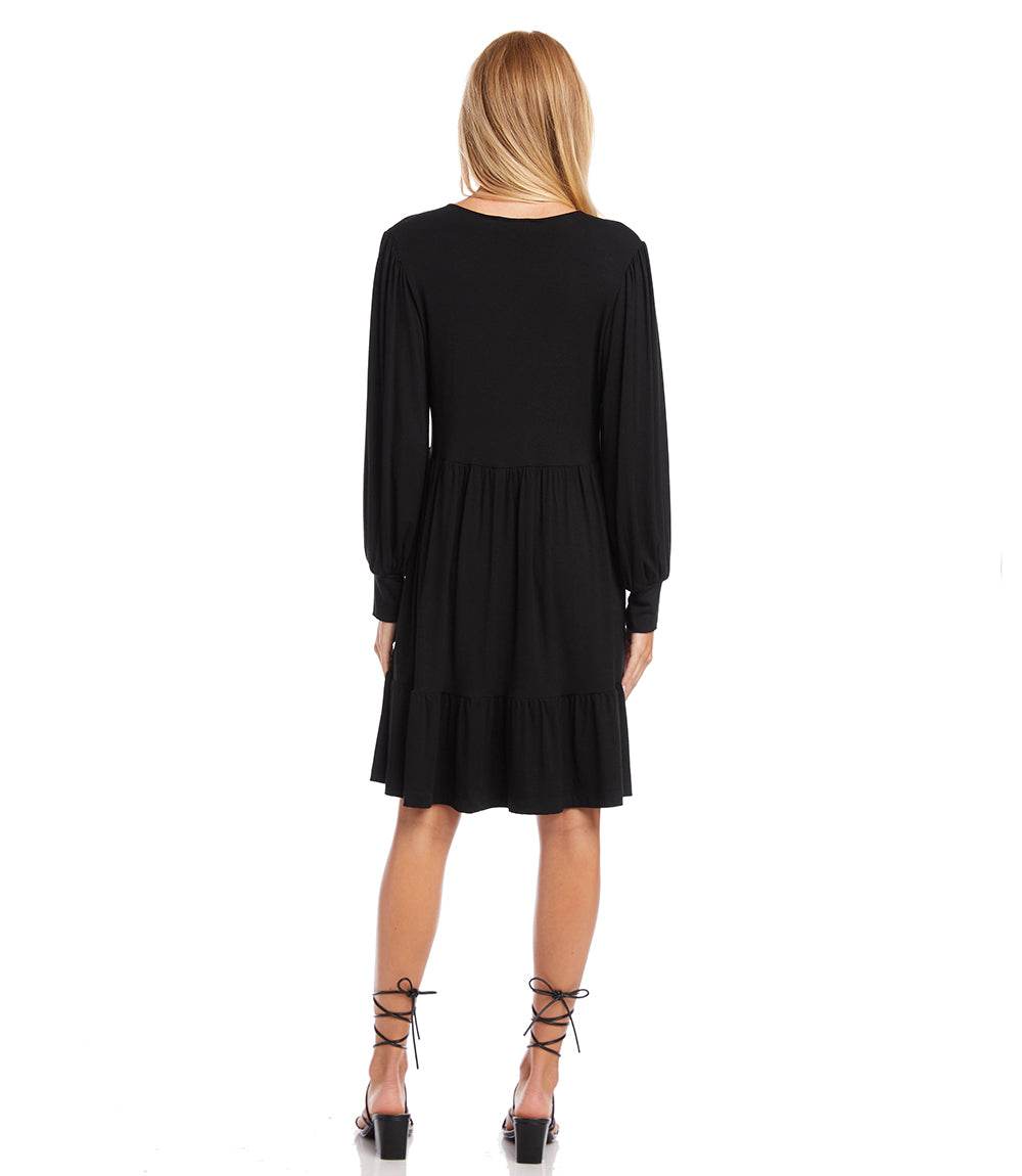 A woman viewed from the back wearing a black long-sleeve dress with a tiered skirt and lace-up high heels against a plain white background.