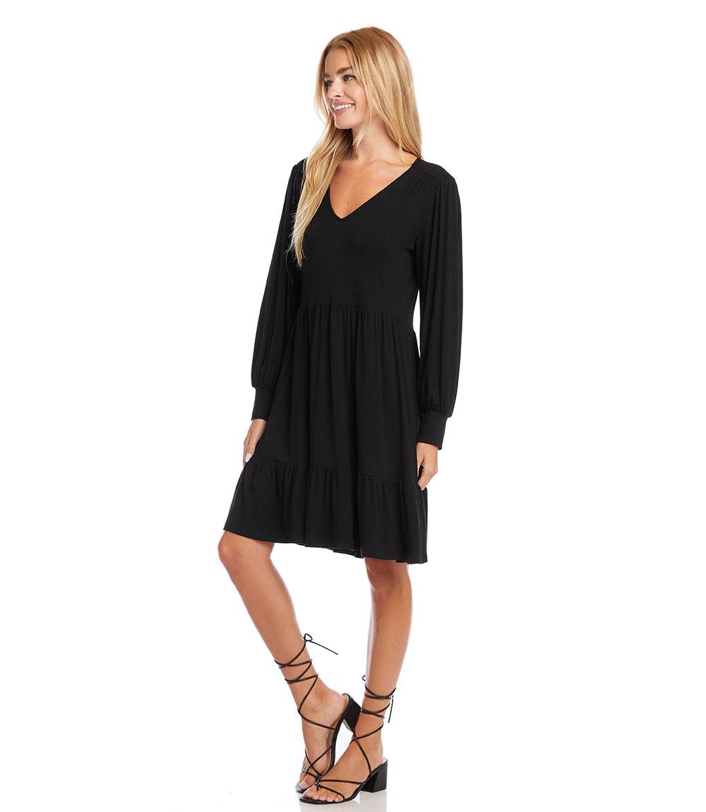 A woman modeling a black long-sleeve V-neck mini dress with a tiered skirt, paired with black lace-up high heels, against a plain white background.