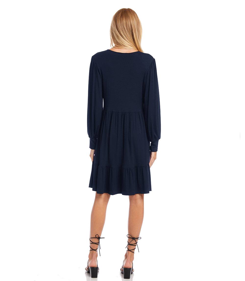 A woman viewed from the back wearing a dark navy blue long-sleeve dress with a tiered skirt, paired with black lace-up high heels, against a plain white background.