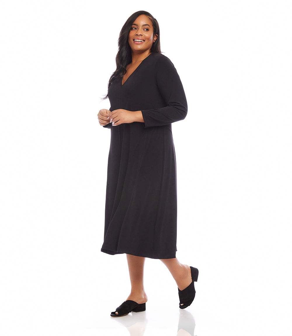 A woman modeling a black long-sleeve midi dress with a V-neckline, standing against a solid white studio background.