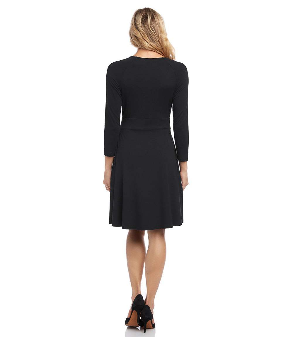 A woman viewed from the back wearing a black long-sleeve knee-length dress with a fitted waist and flared skirt, paired with black heels, against a plain white background.