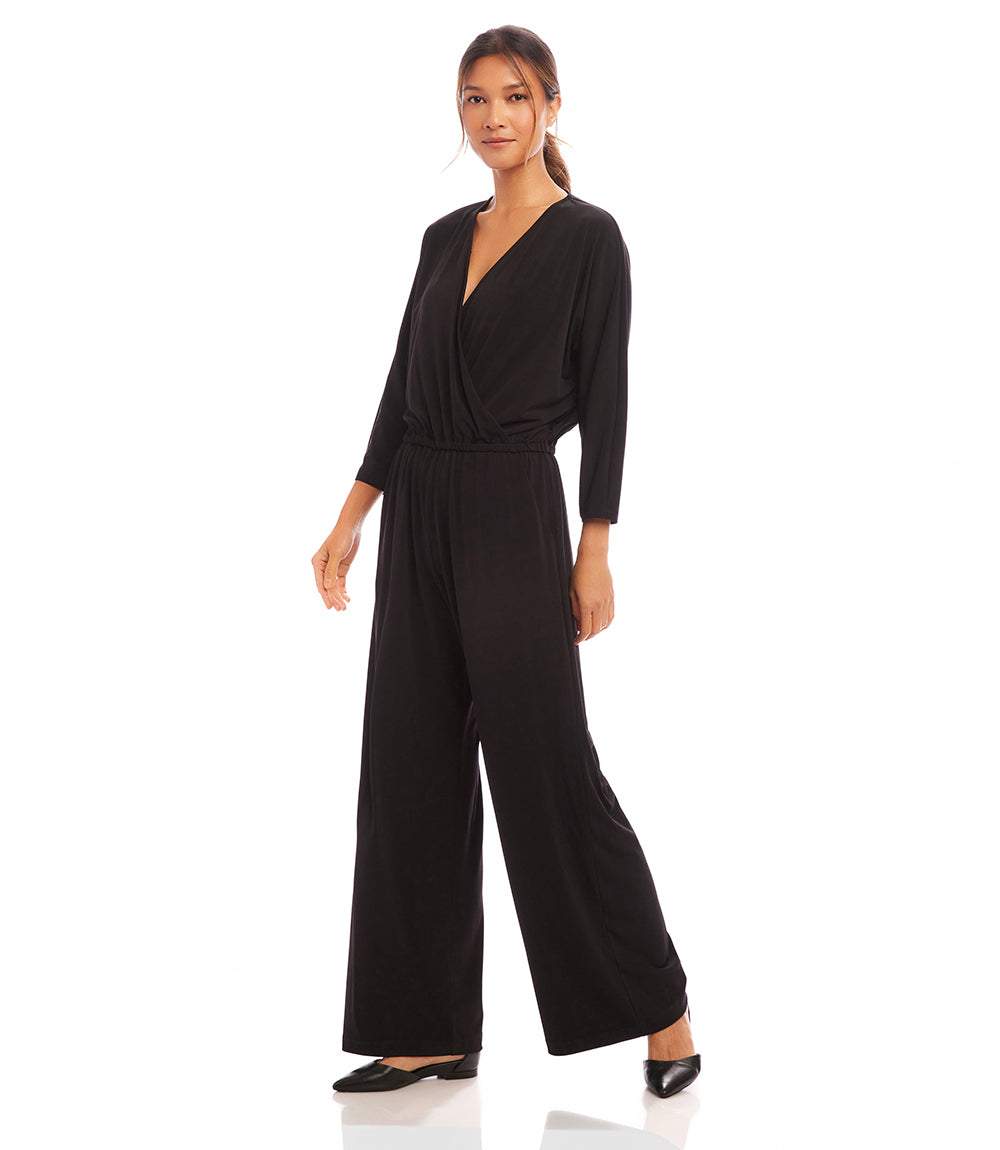 A woman modeling a black wrap-style jumpsuit with three-quarter sleeves and wide legs, standing against a plain white studio background.