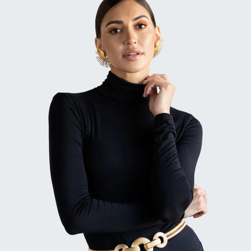 A woman modeling a black long-sleeve mock neck top and matching black trousers, styled with a gold chain belt and gold earrings, against a plain light gray background.