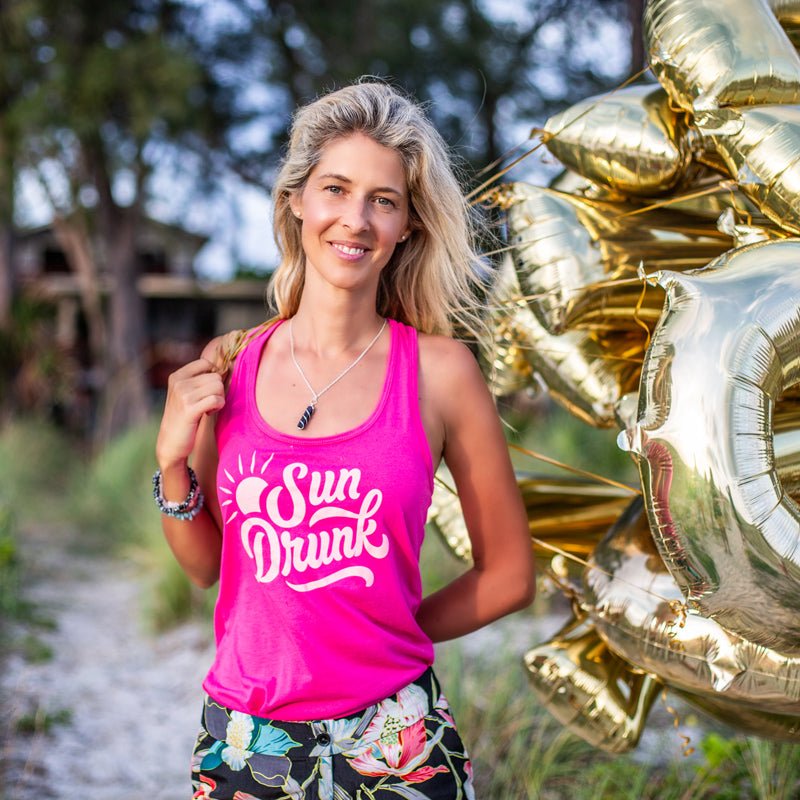 Woman wearing a pink 'Sun Drunk' tank top with gold balloons in the background.