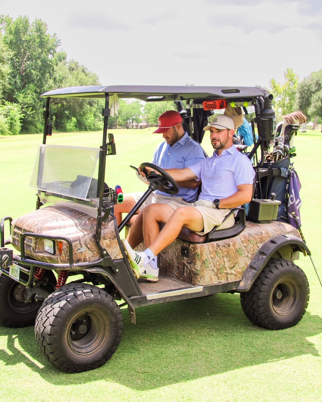 Two men sitting in a camouflage-patterned golf cart on a golf course, with trees and grass in the background.