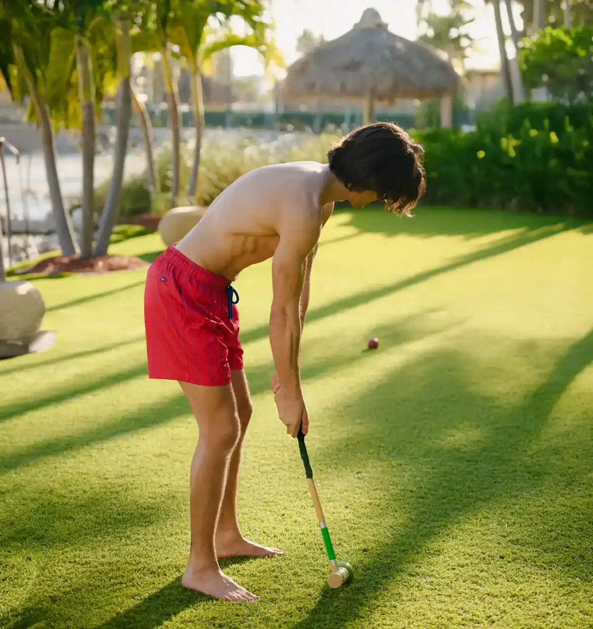 Man in red shorts playing mini golf on a sunny day with palm trees in the background