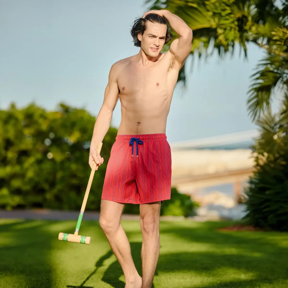 Man in red swim shorts holding a croquet mallet on a grassy area with palm trees in the background