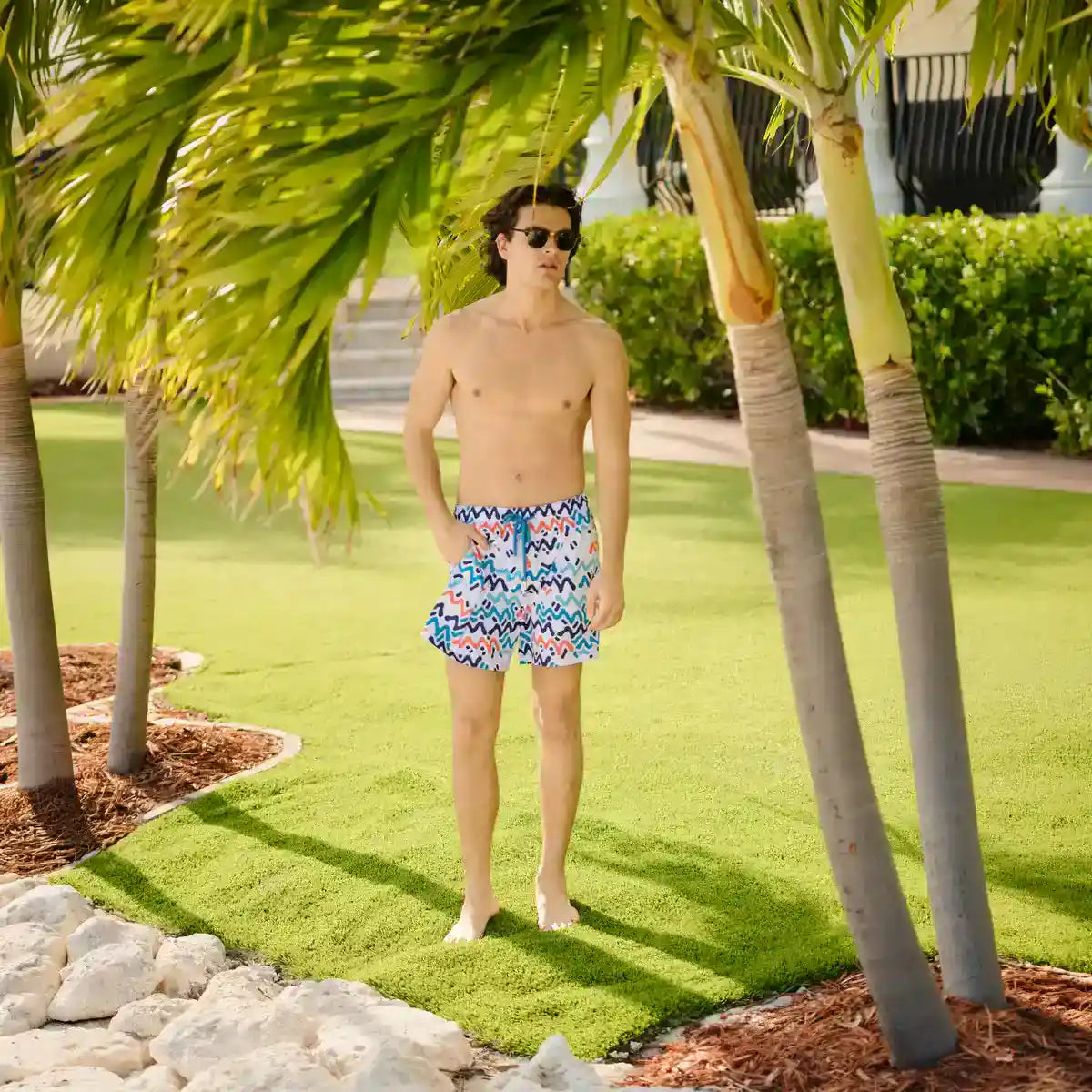 Man in colorful swim shorts standing under palm trees on a sunny day.