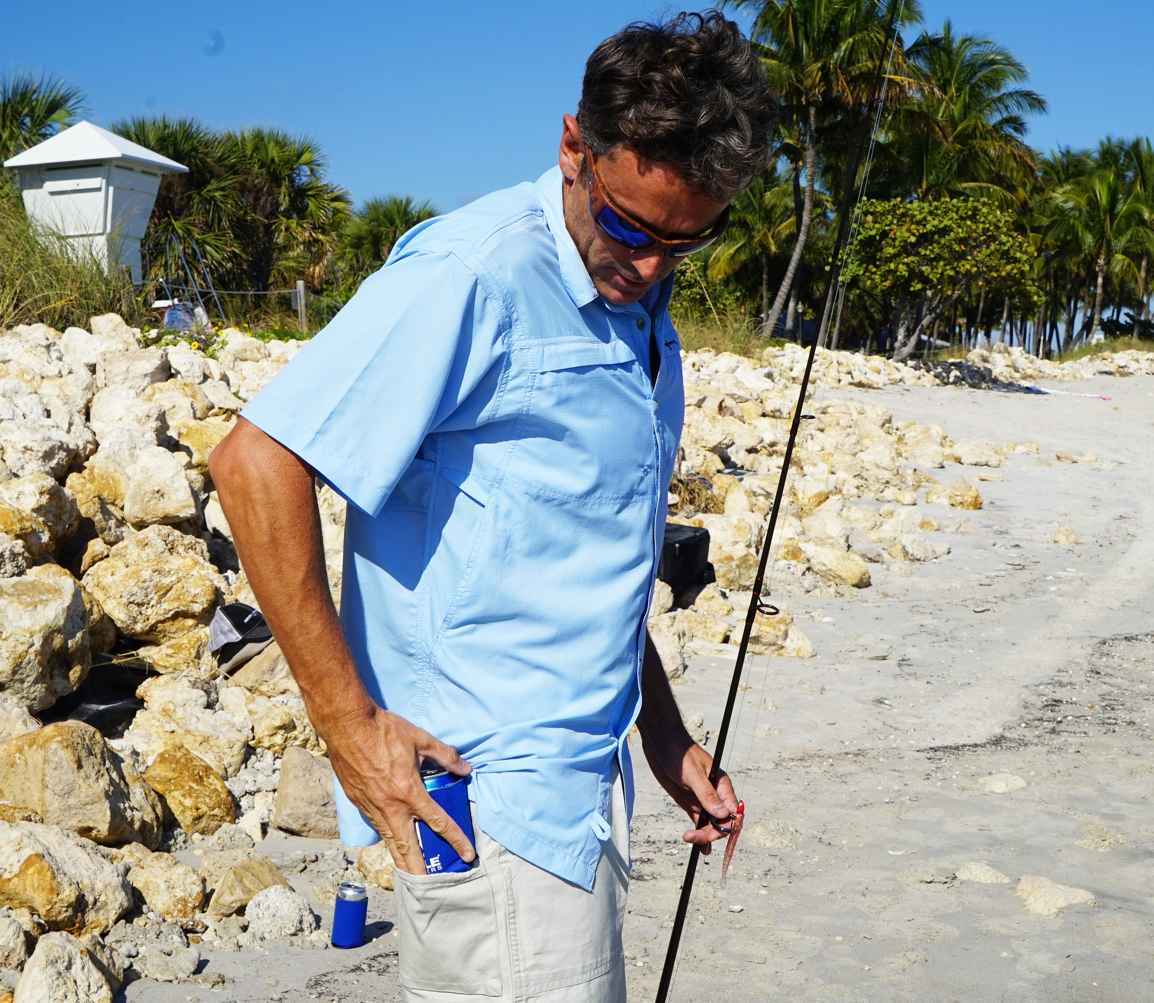 Man in light blue shirt holding a fishing rod on a beach with rocks and palm trees in the background.