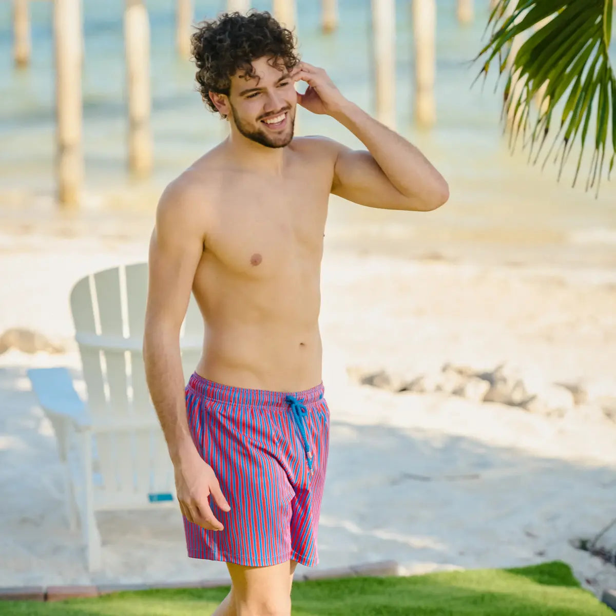 Man wearing colorful swim shorts standing by a beach with palm trees and lounge chairs.
