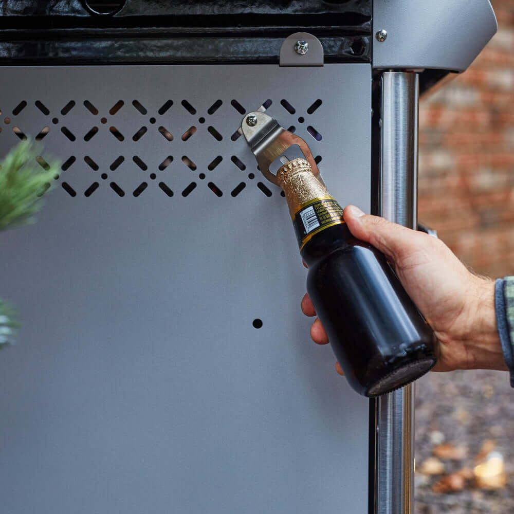 Person using a bottle opener on a beer bottle in front of a grill.