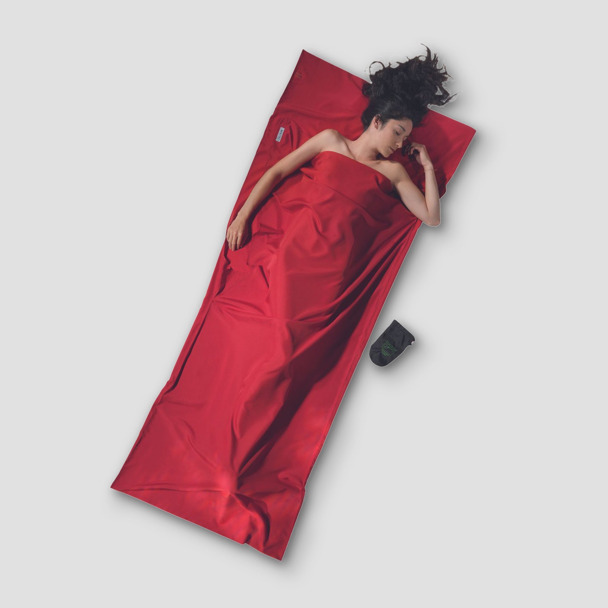 Person lying on a red sleeping bag with a pillow and device on a light gray background