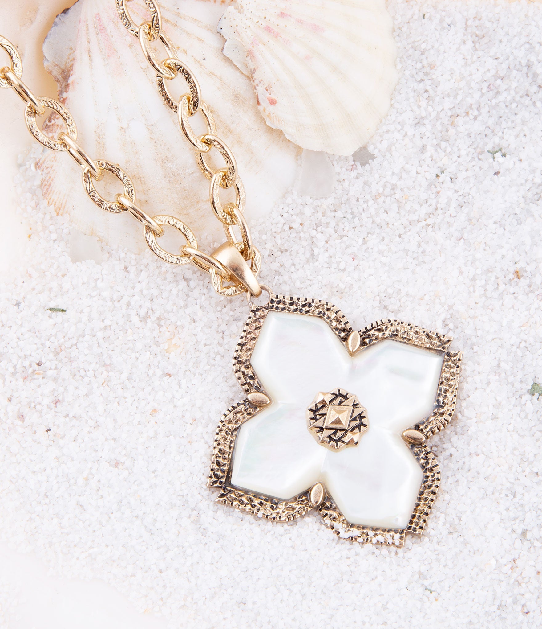 Gold necklace with a heart-shaped pendant featuring a white flower design on a sandy background.