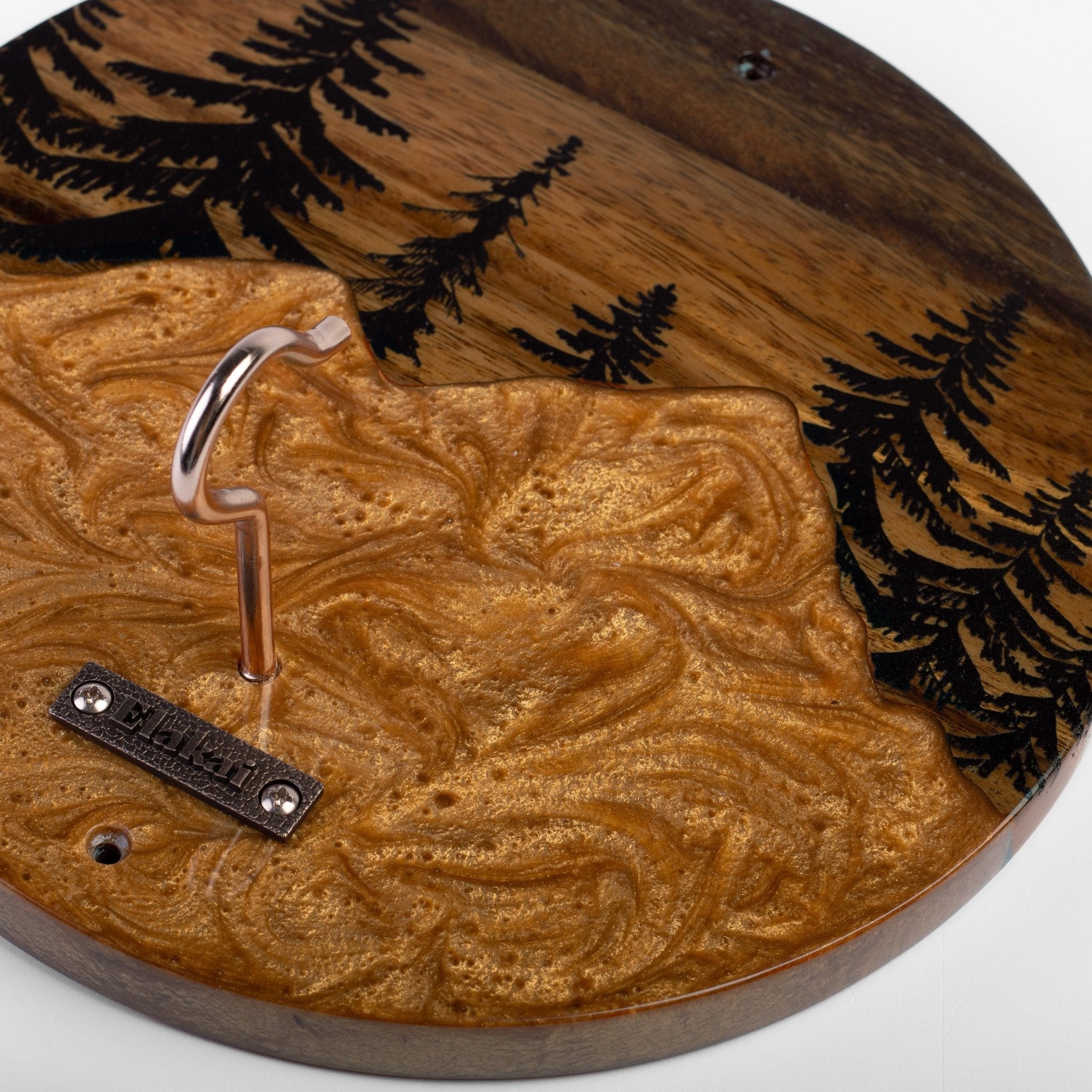 A close-up of a round wooden wall hook with a mountain-shaped textured gold-toned front, a dark wood top with black pine tree engravings, and a metal hook mounted on the side.