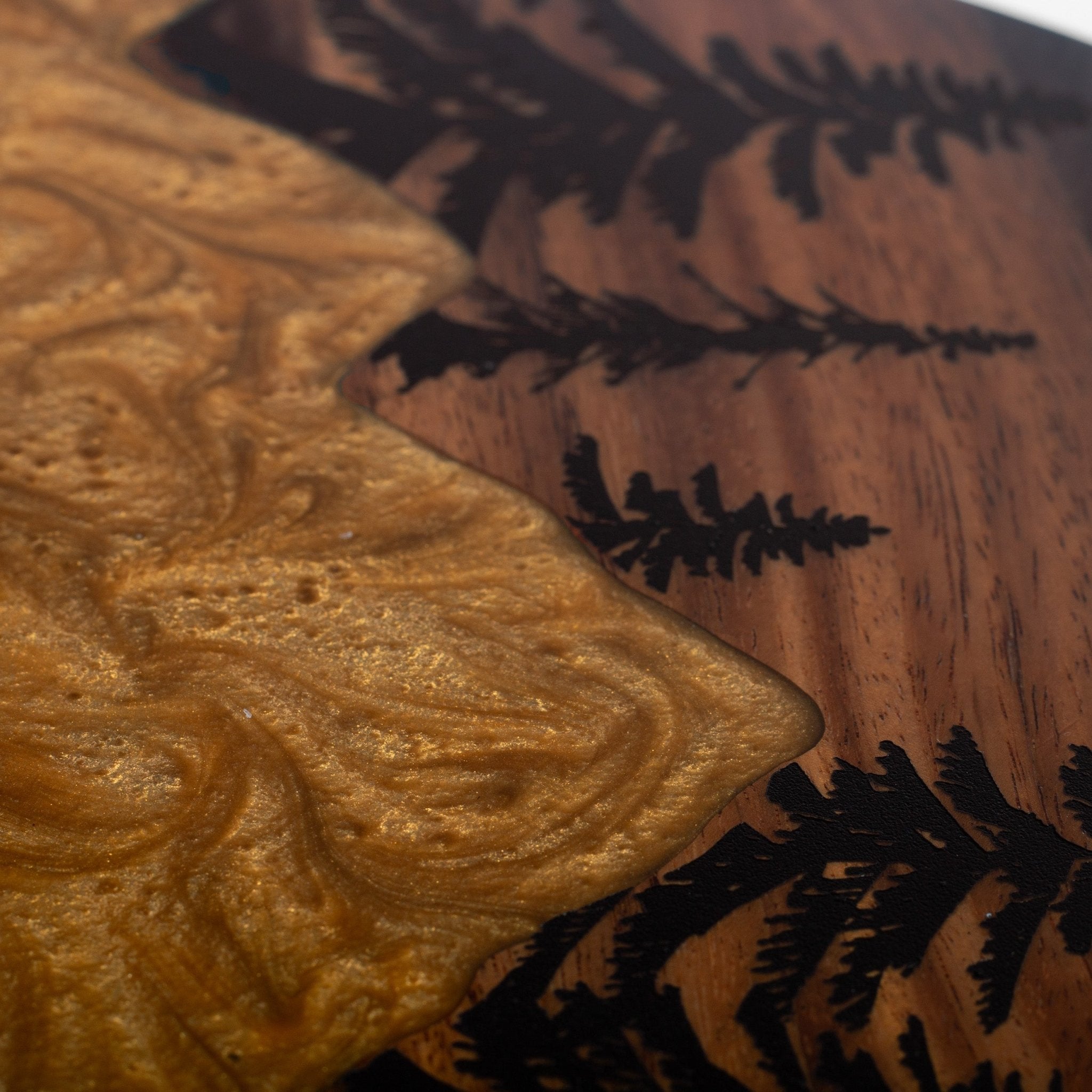 Close-up of a decorative wooden surface with a wavy metallic gold resin inlay and dark burned or inlaid tree-branch silhouettes on a brown wood base.