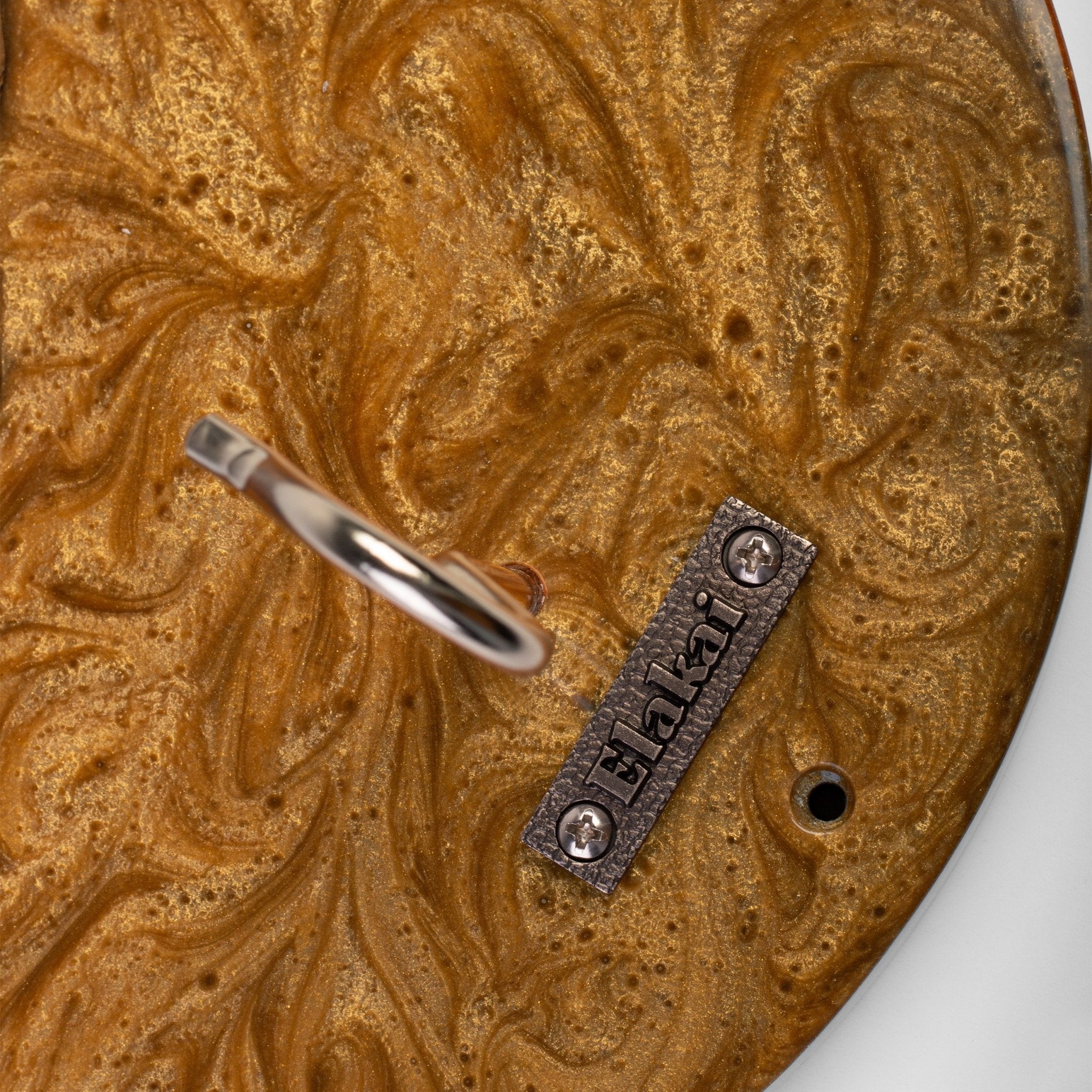 Close-up of a round decorative object with a marbled gold and brown surface, featuring a silver metal ring and a small engraved metal plaque reading 'Hakl'.