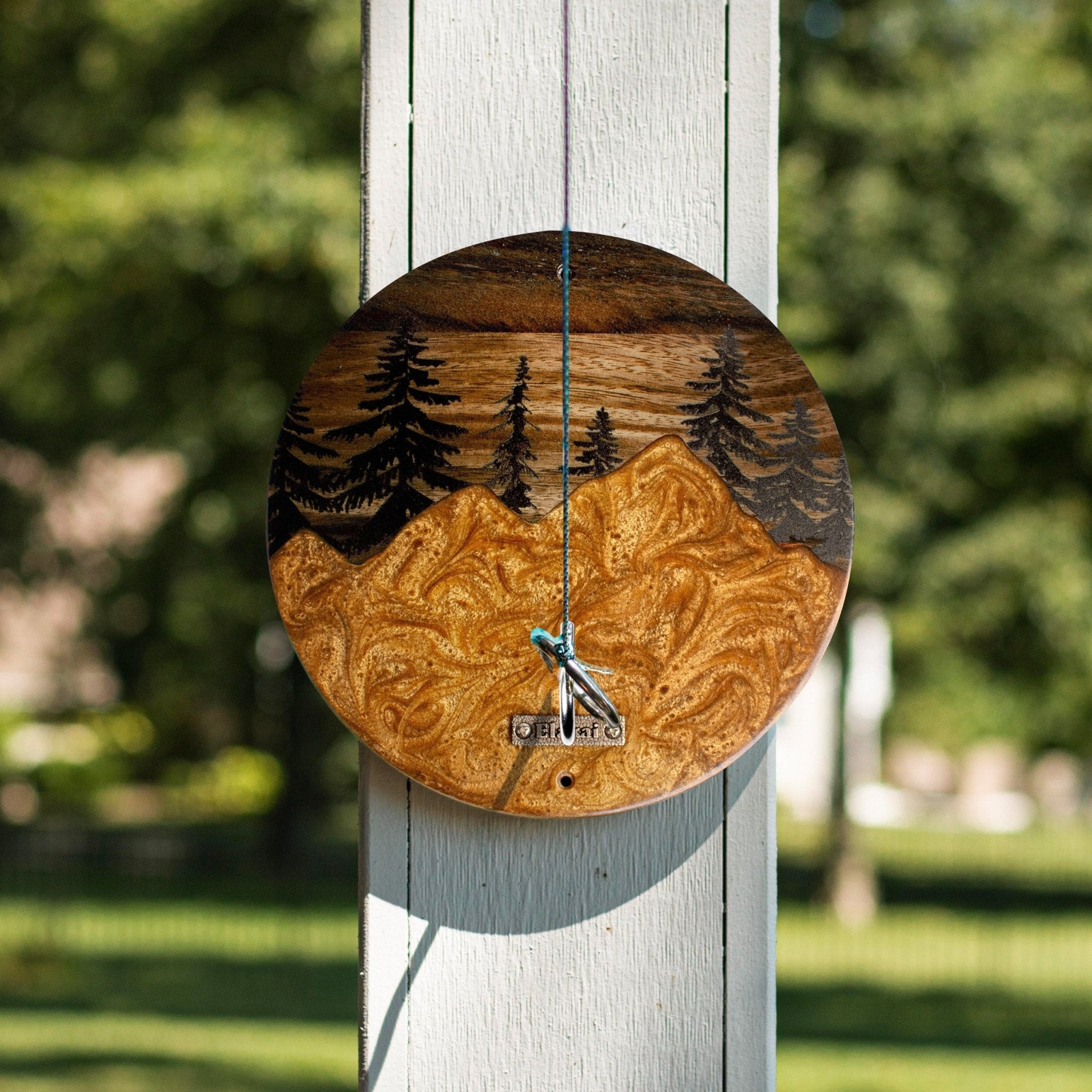 A round wooden wall clock with a mountain and pine tree design, hanging on a white wooden post outdoors with a blurred green background.