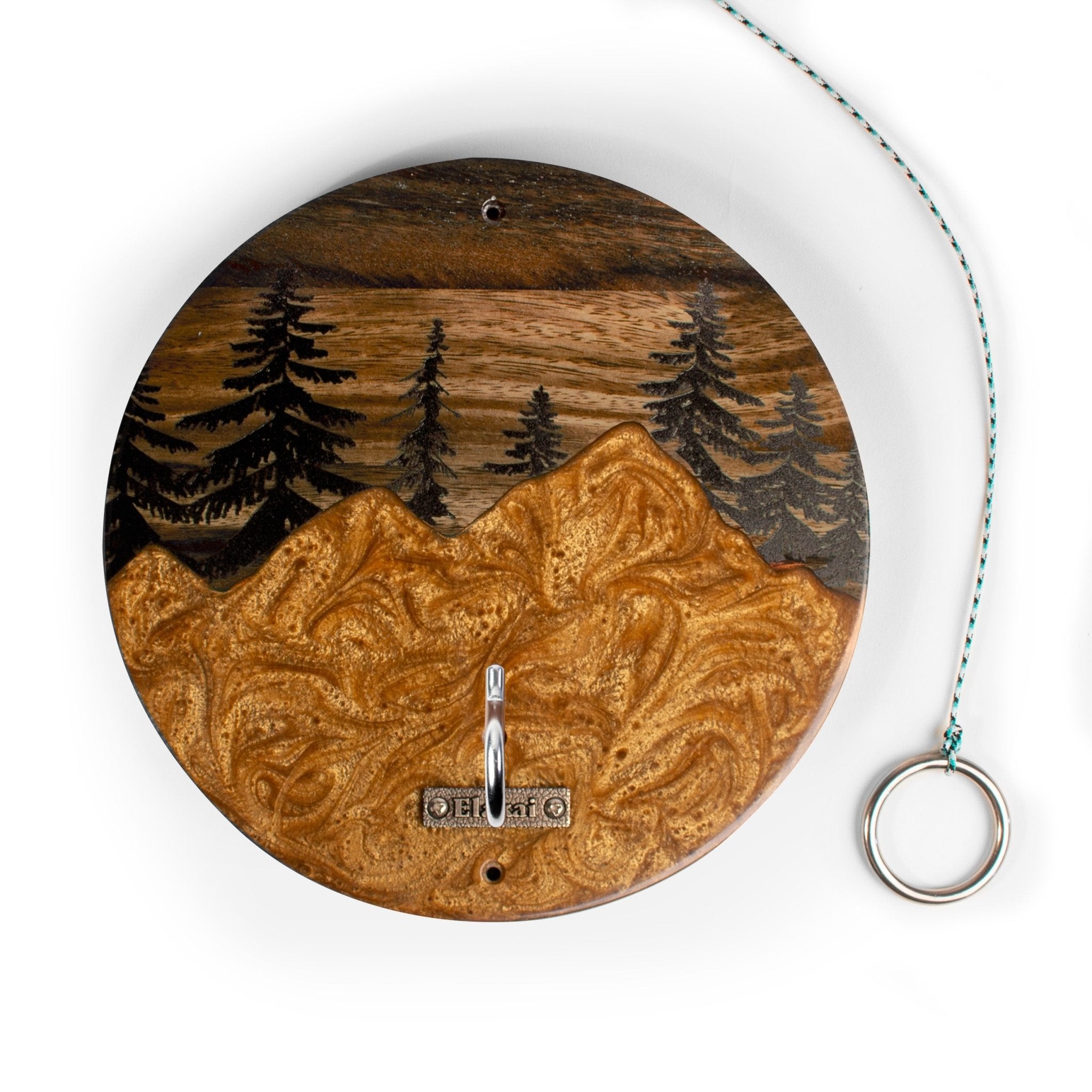 A round wooden wall clock featuring a carved mountain and pine tree landscape design. The clock has a silver metal hanging hook at the bottom and a teal-and-white string with a silver ring pull on the right side, set against a plain white background.