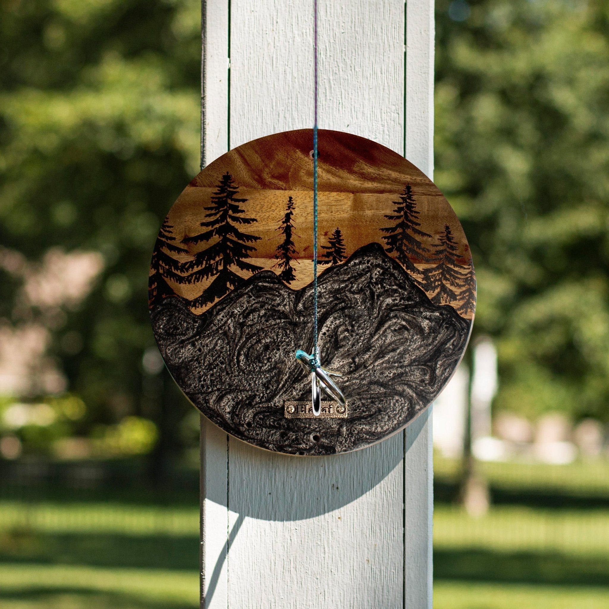A round wooden wall clock with an engraved mountain and pine tree landscape design, hanging on a white wooden post outdoors with a blurred green garden background.