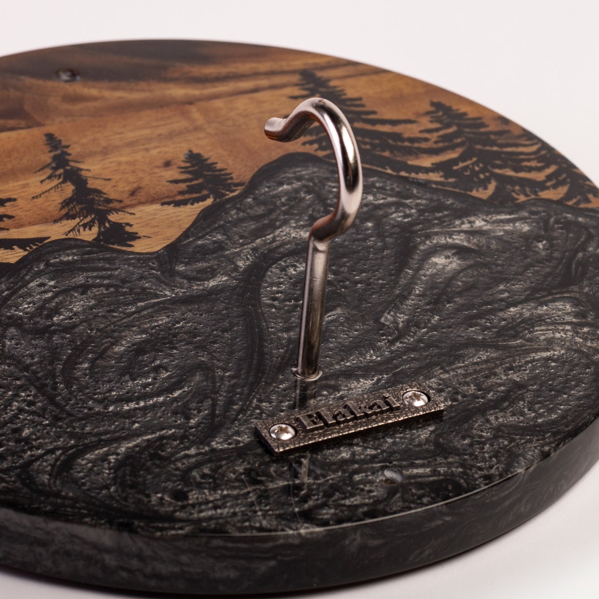 A close-up of a round wooden wall hook with a mountain and forest design. The hook is mounted on a dark, textured faux-snow mountain face, with the wood and tree engraving visible in the background.
