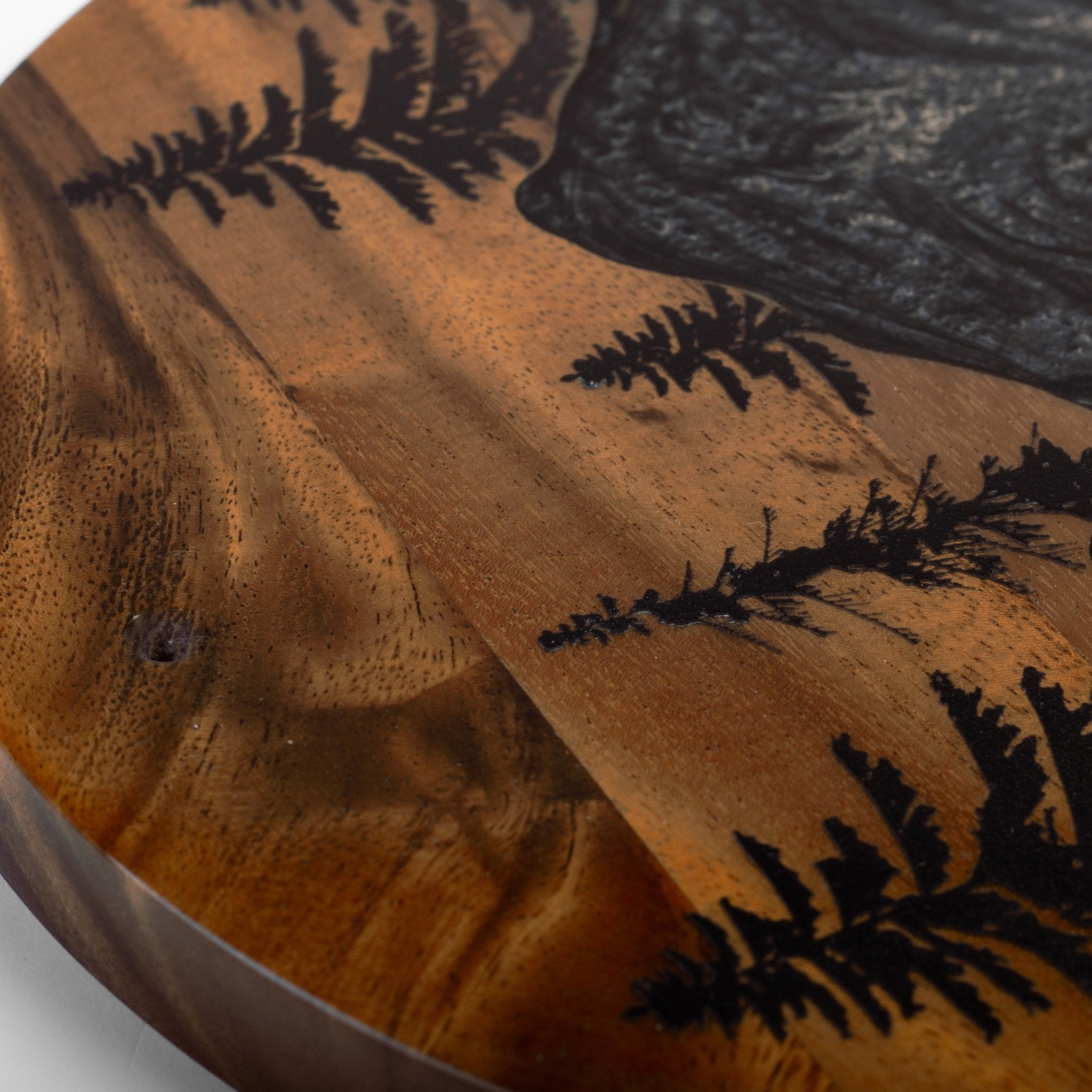 Close-up of a round wooden wall plaque featuring a natural brown wood grain background with black laser-engraved fern patterns and a dark textured inlay in the upper right.