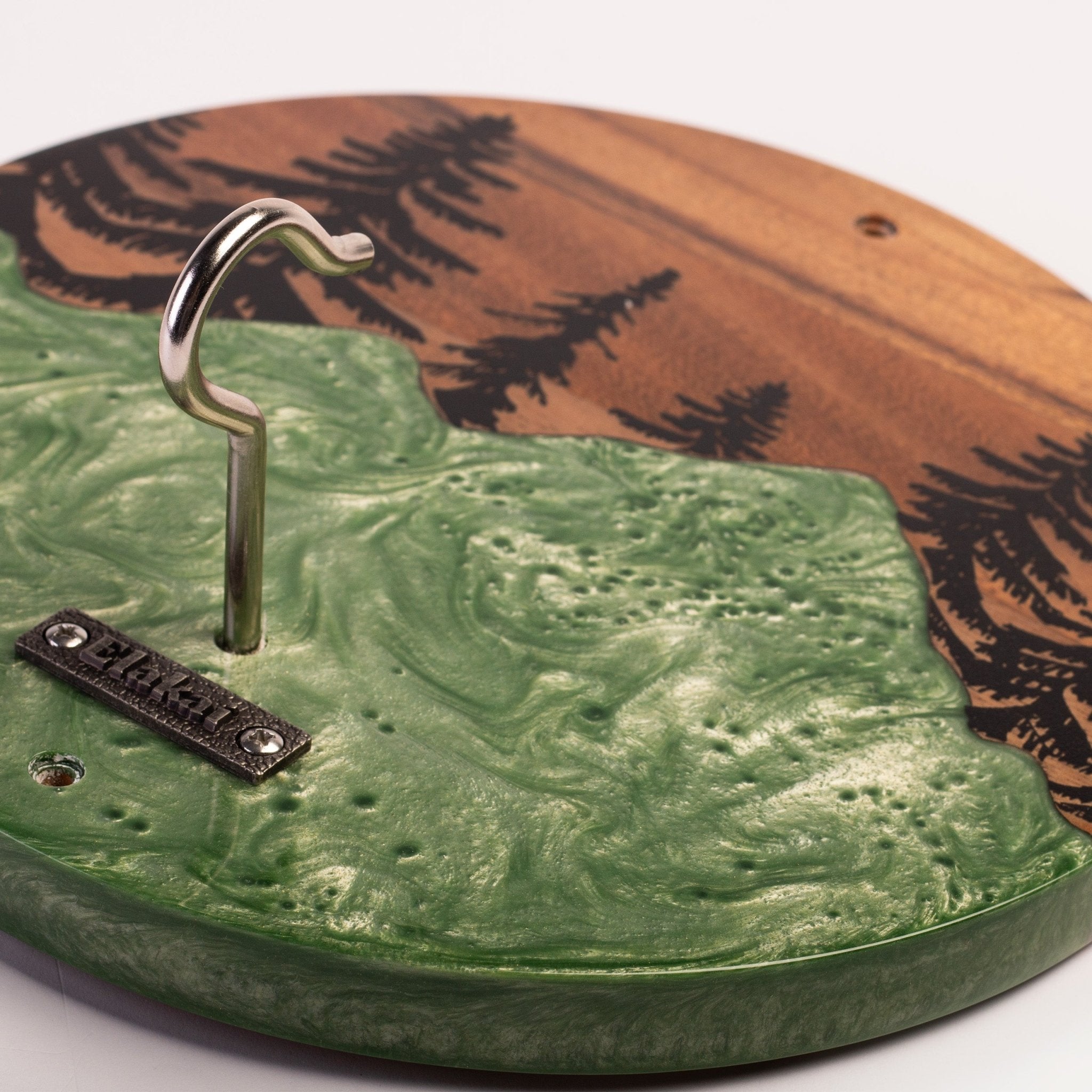A close-up of a round wooden pegboard with a green textured lower section and a brown upper section featuring black tree silhouettes. A silver metal hook is attached on the left, along with a small black brand plaque that reads 'BARNES'.