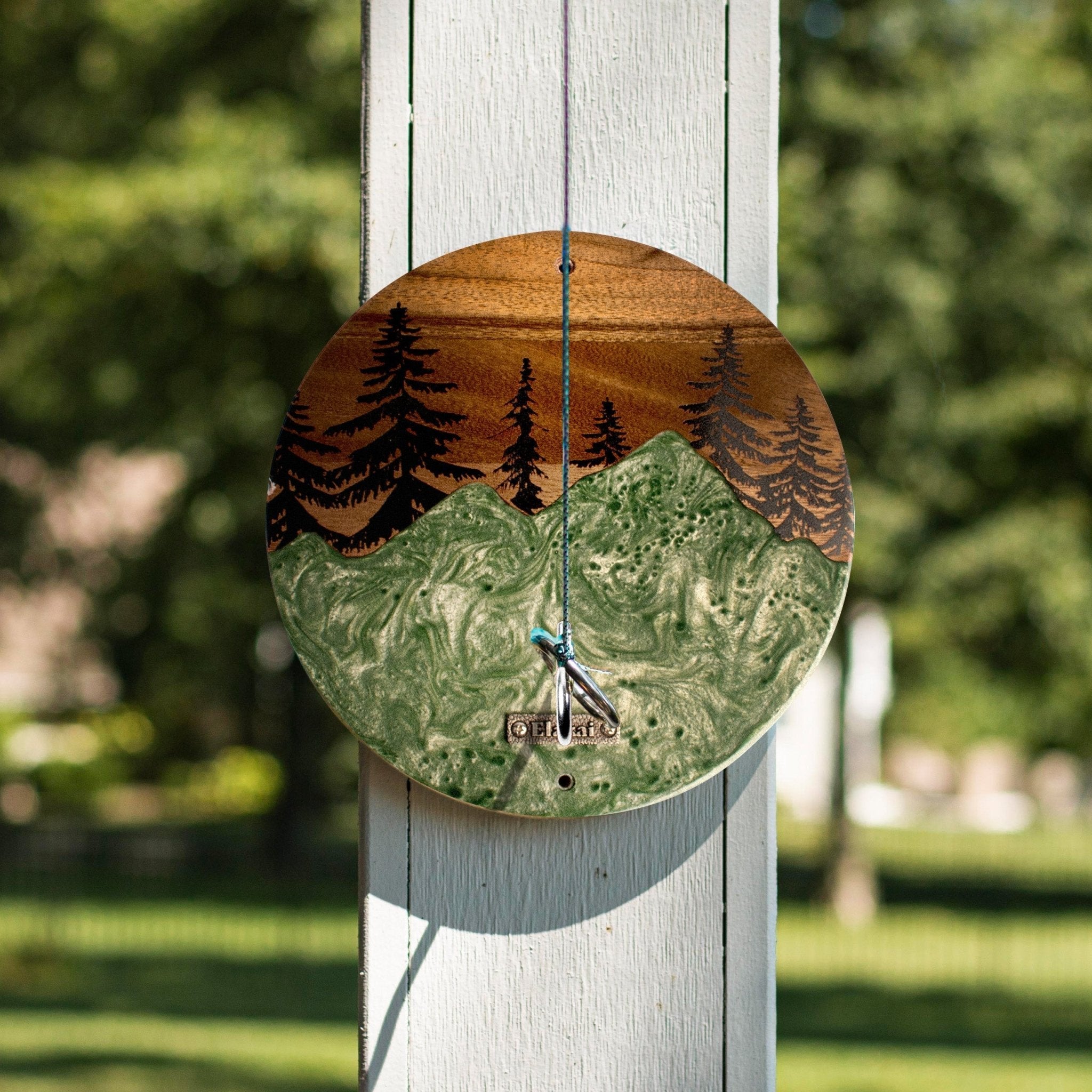 A round wooden wall clock with a mountain and pine tree landscape design, hanging on a white wooden post outdoors with a blurred green garden background.