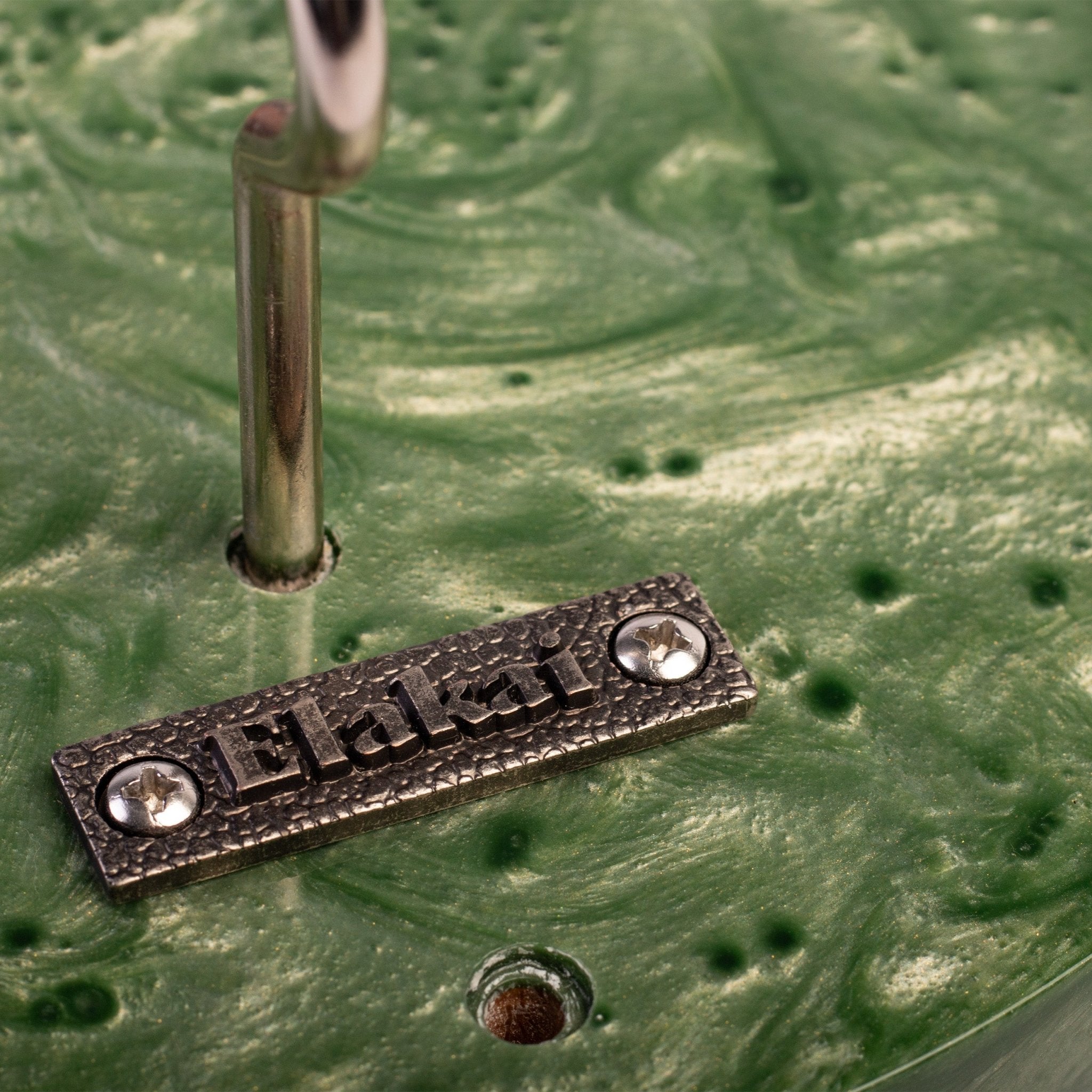 Close-up of a textured green circular surface with a small rectangular metal plate engraved with the word "Ehat" and two silver screws, alongside a vertical metallic rod.