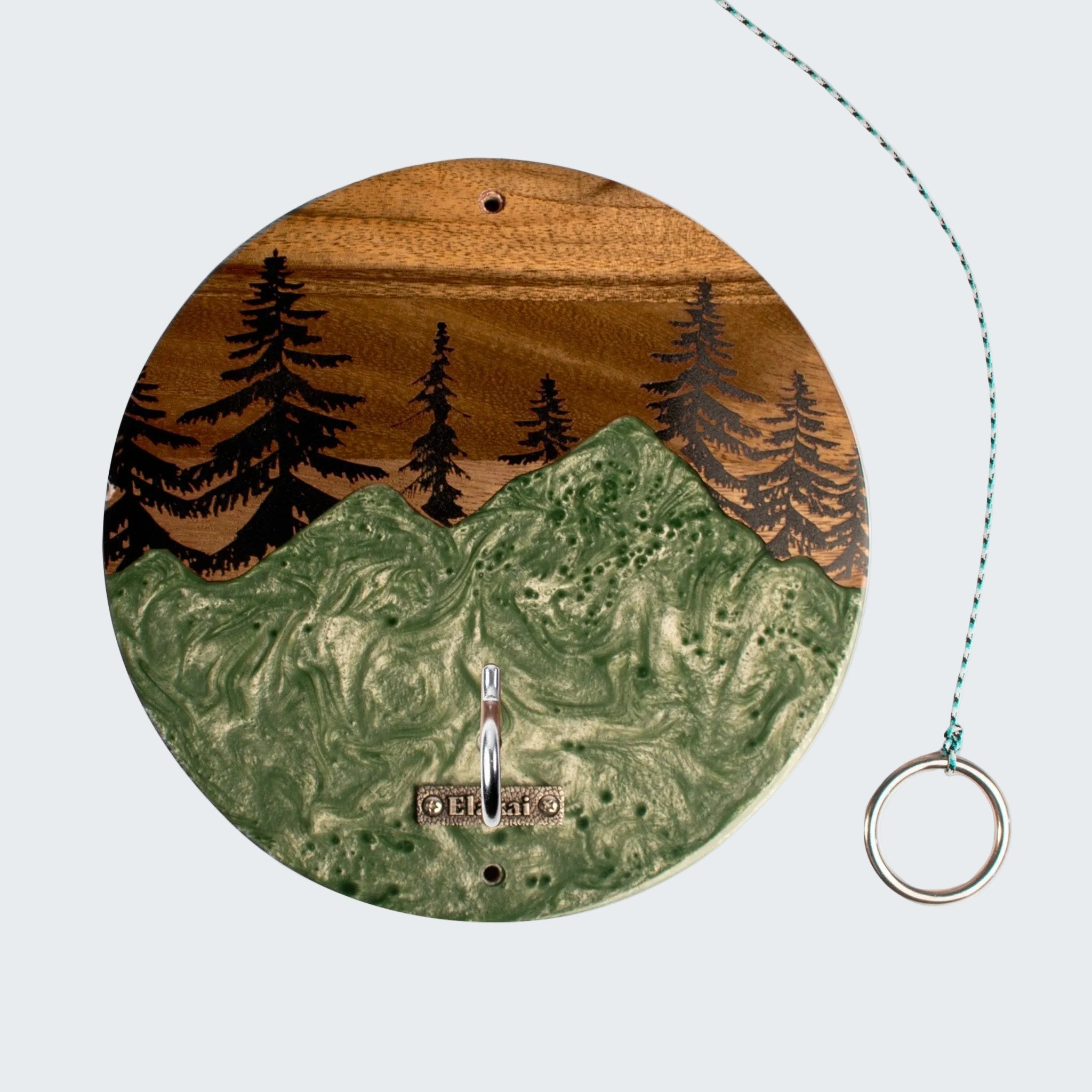 A circular wooden wall clock featuring an engraved landscape design with green mountains, black pine trees, and a brown sky. The clock has a silver metal hook at the bottom and a teal-and-white string with a silver ring pull on the right, set against a plain white background.