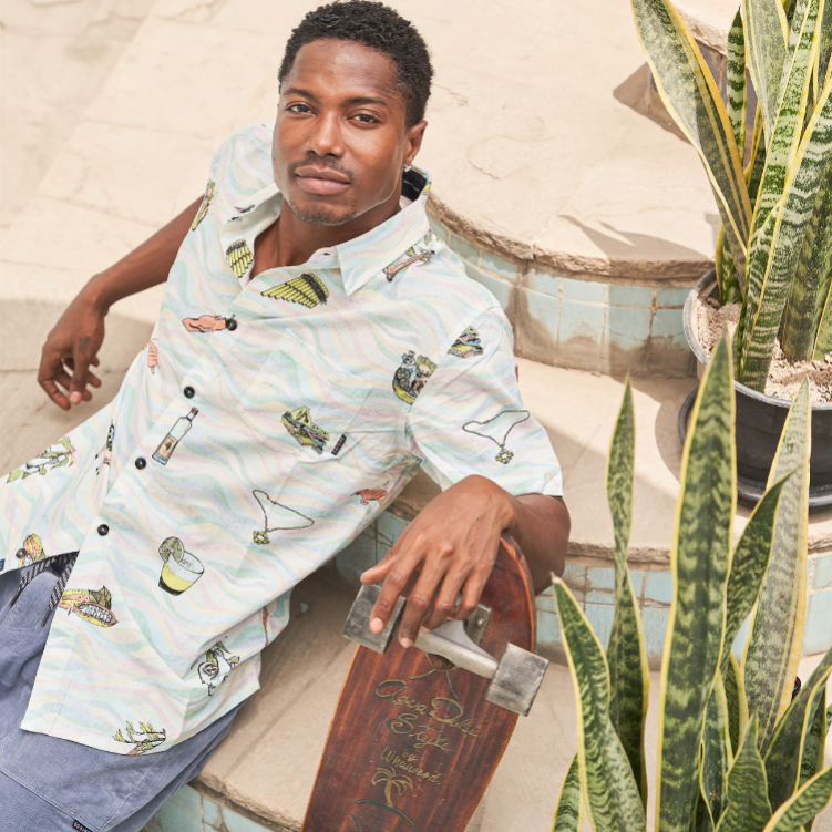 A man wearing a white short-sleeve button-up shirt with a tropical cocktail print, sitting on tiled steps next to a skateboard and potted plants.