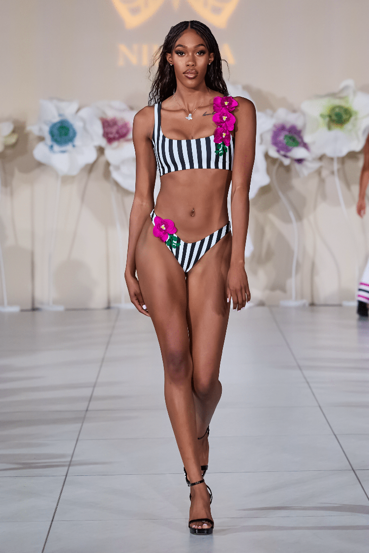 Model walking on a runway wearing a black and white striped bikini with pink floral accents.