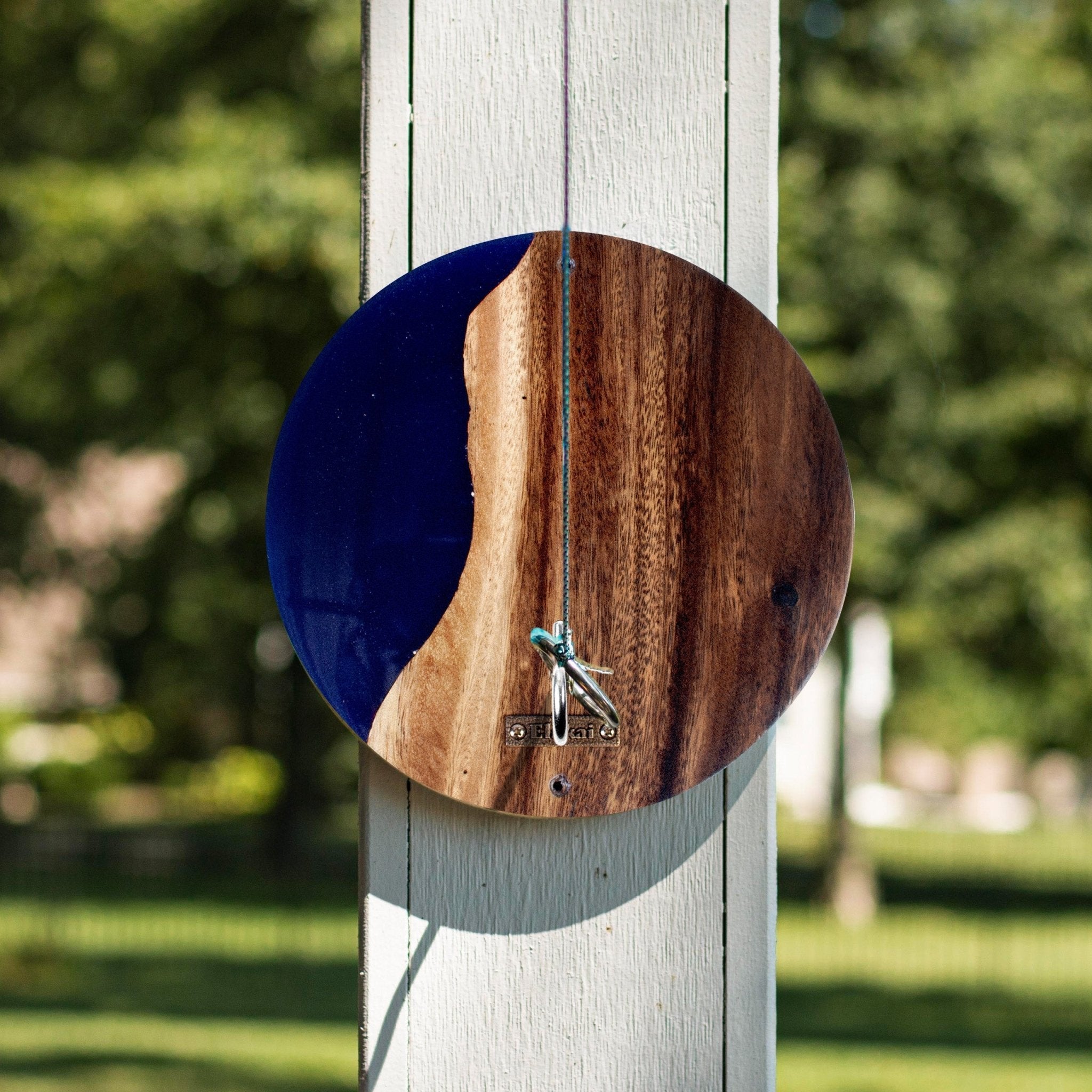 A circular wall clock with a split design, featuring a dark blue left half and a natural brown wood grain right half, hanging on a white wooden post in an outdoor garden setting.