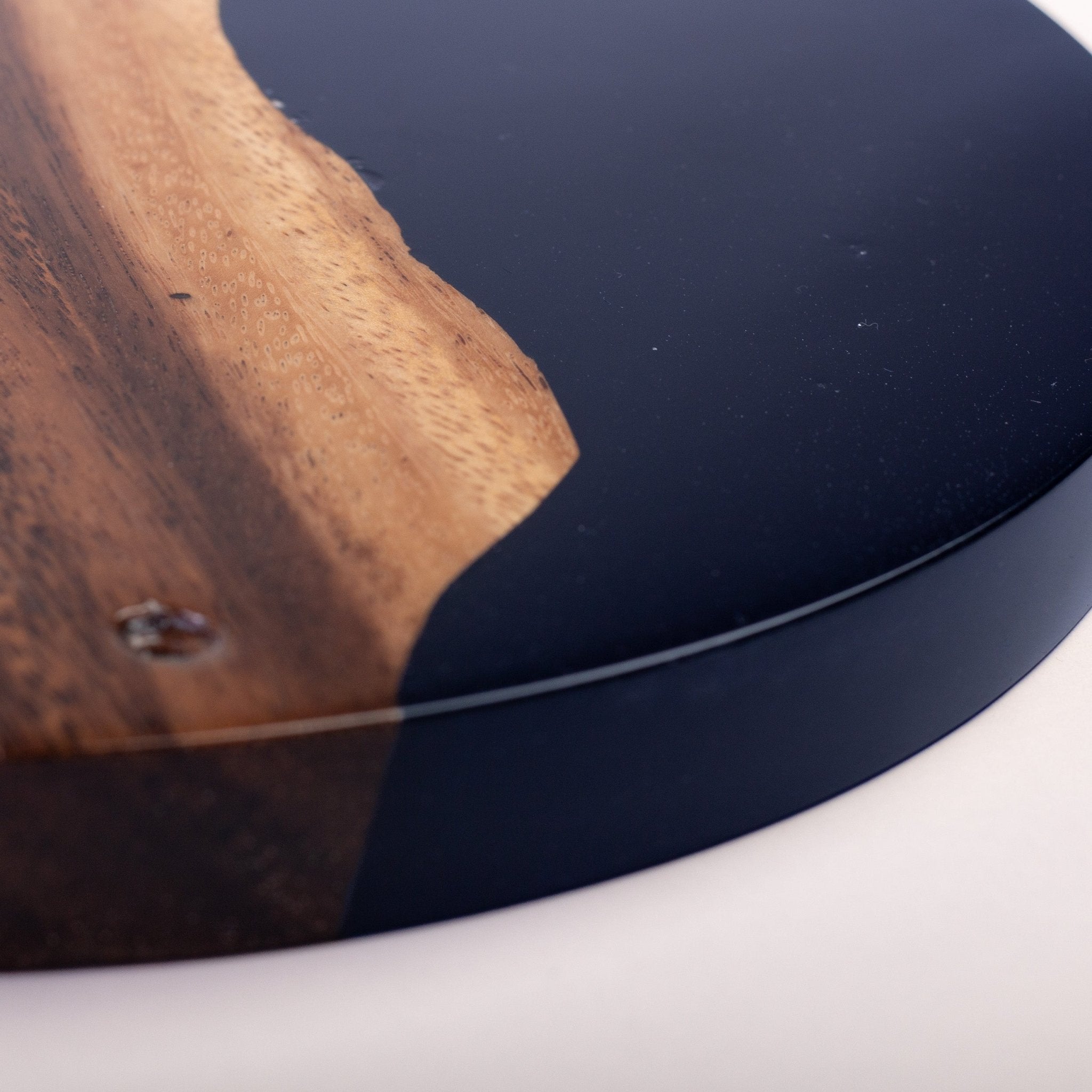 Close-up of a round wooden board or tray with a glossy black resin inlay and a natural live-edge wood pattern, shown on a white surface.
