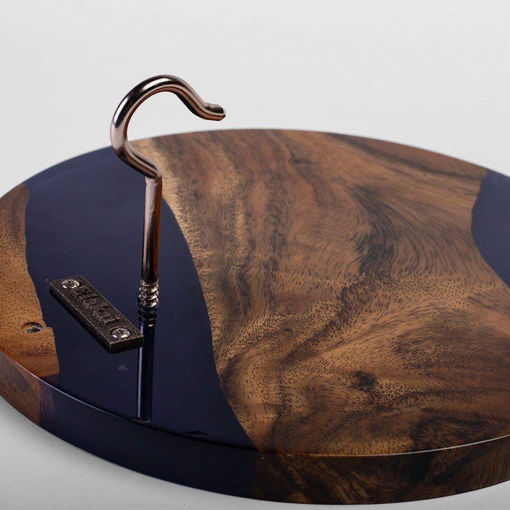 A close-up of a round wooden serving board or tray with a dark polished resin edge, a natural wood grain center, and a metal hook hanging from a stand.