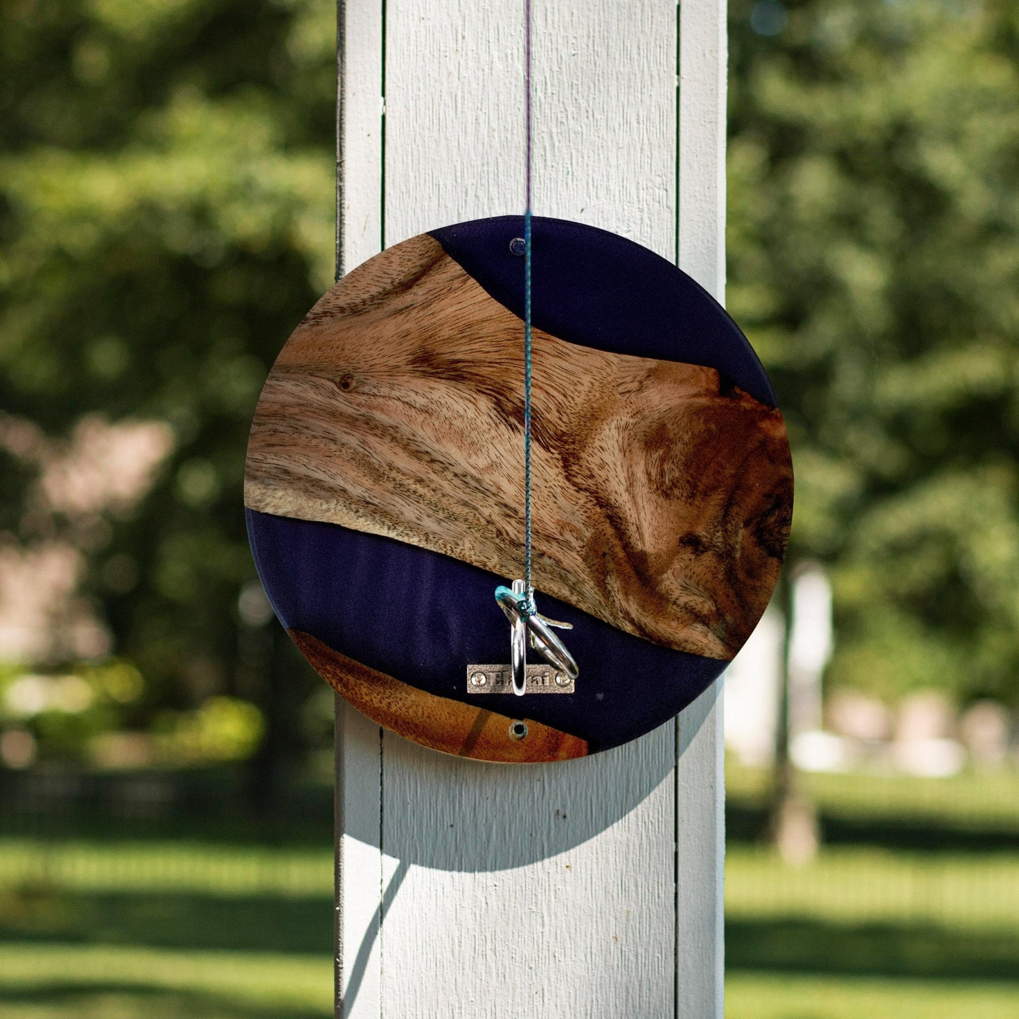 A circular wooden wall hanging with a natural wood grain pattern and dark navy blue sections, suspended by a string from a white wooden post in an outdoor garden setting.