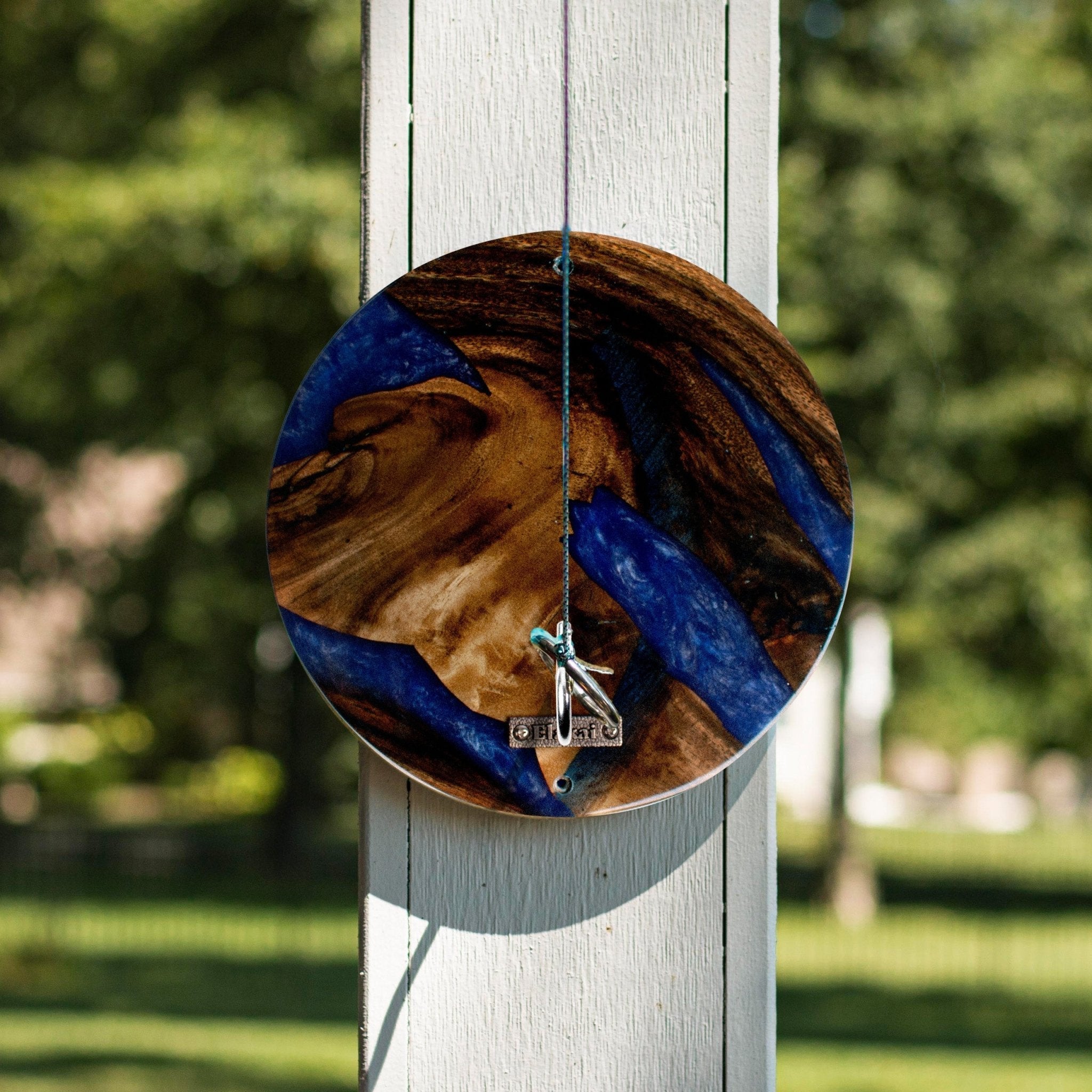 A round wooden wall hanging with a blue and brown resin pattern, suspended by a string from a white wooden post in an outdoor garden setting.