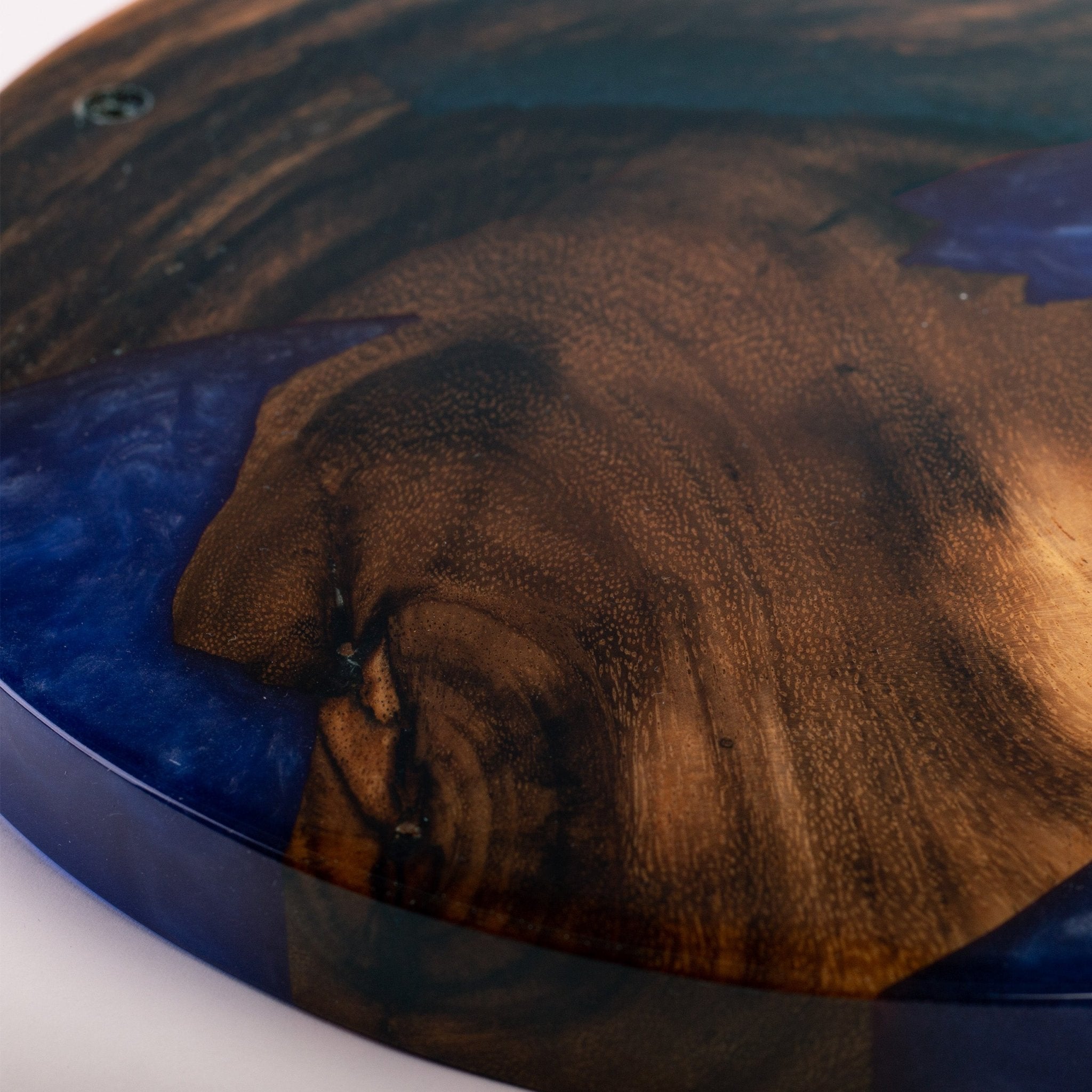 Close-up of a round wooden plaque or coaster with a glossy finish, featuring a swirling pattern of dark brown wood grain and deep blue resin inlay.