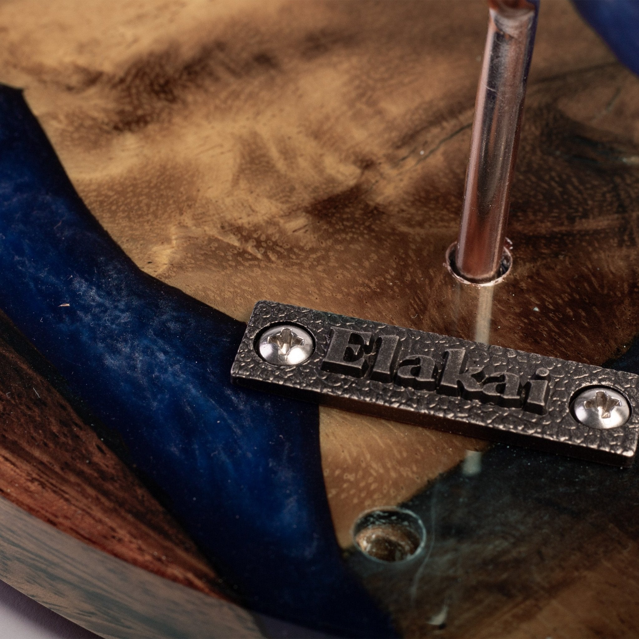 Close-up of a wooden item with a dark blue resin inlay and a metal plate engraved with the brand name 'Elakai'.