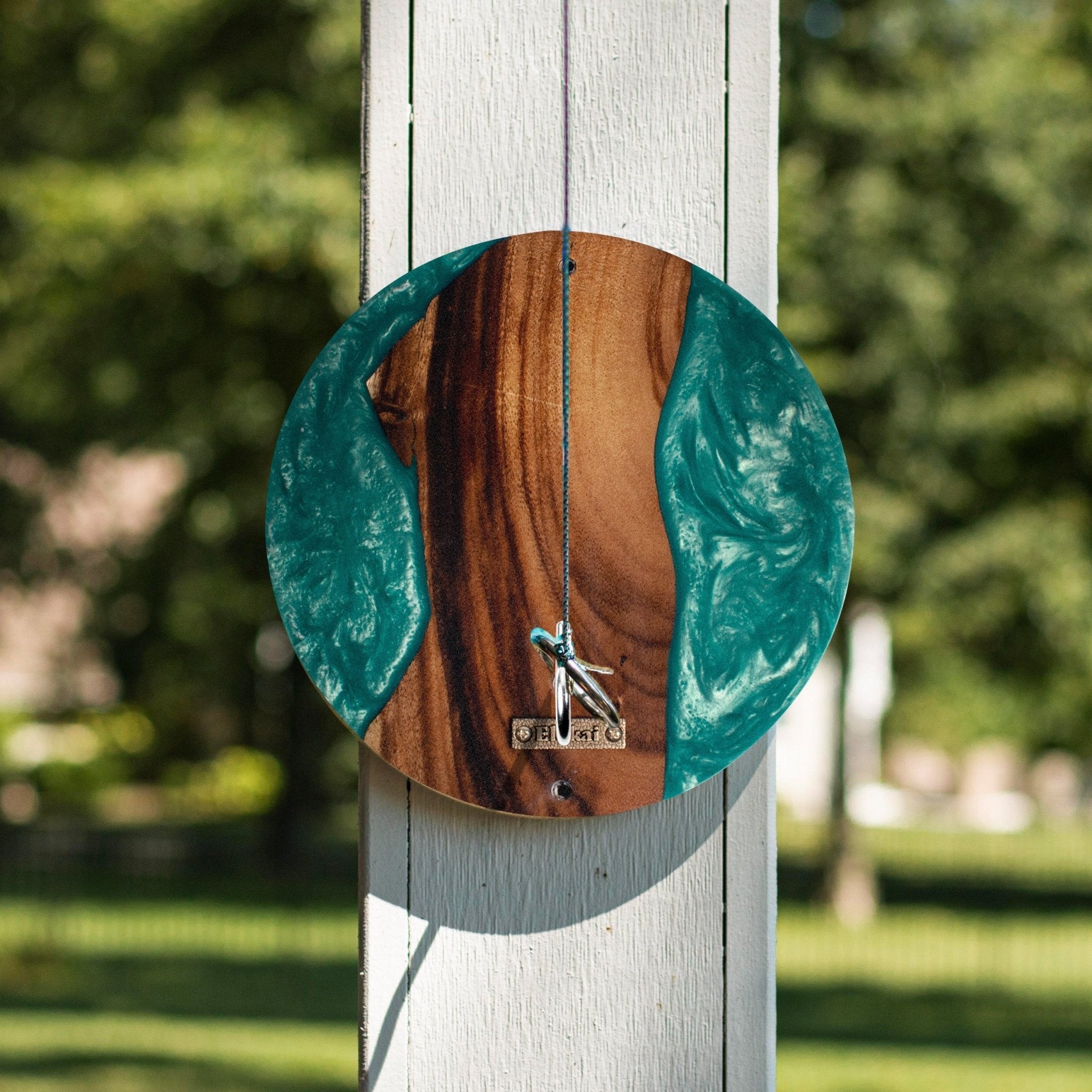 A circular wooden wall clock with a river-like design, featuring a brown wood grain center and teal resin sections on the sides. A small metal figure and 'OCTO' text are visible on the clock face.