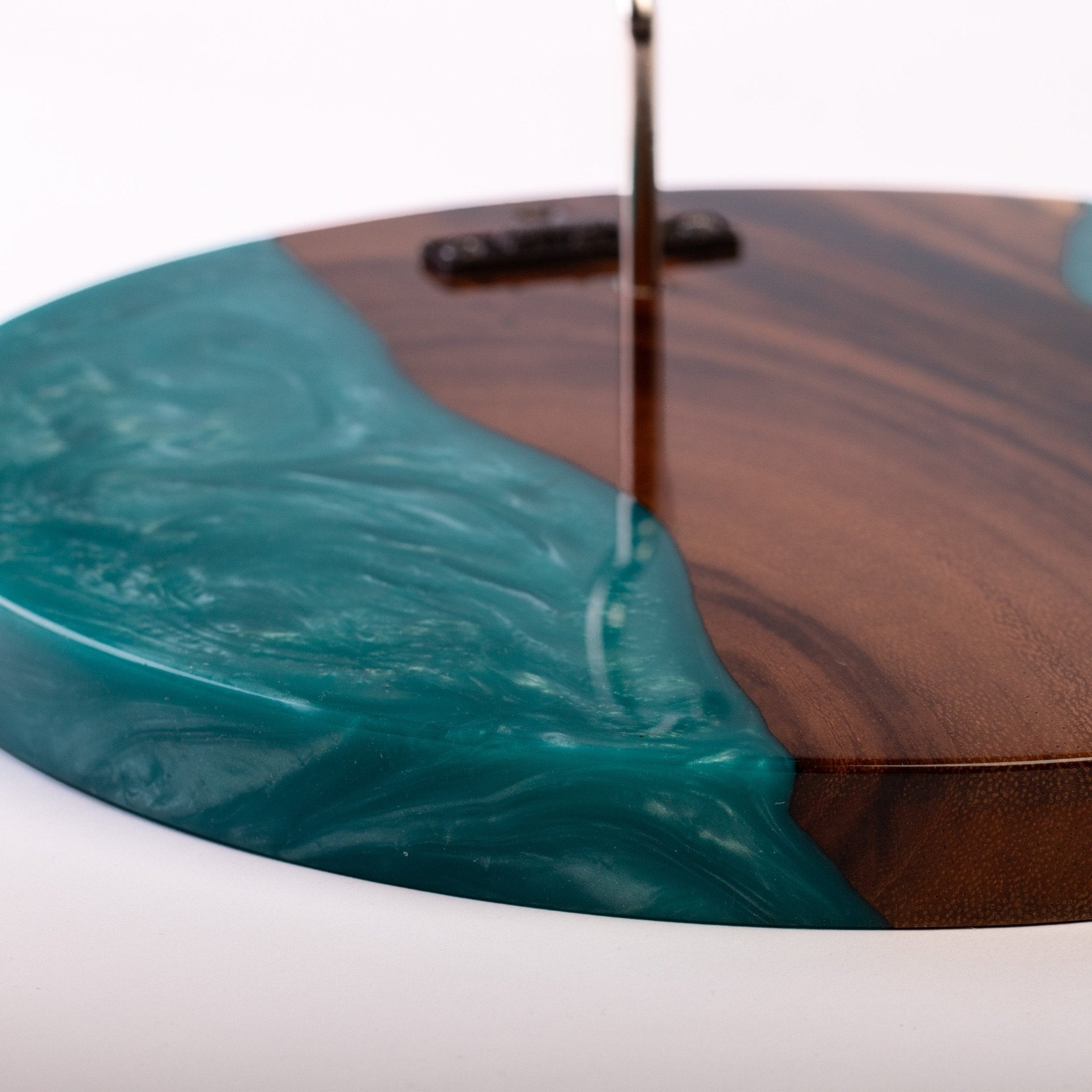 A close-up of a round wooden tray or plate with a glossy teal resin swirl flowing across the surface, photographed on a white background.