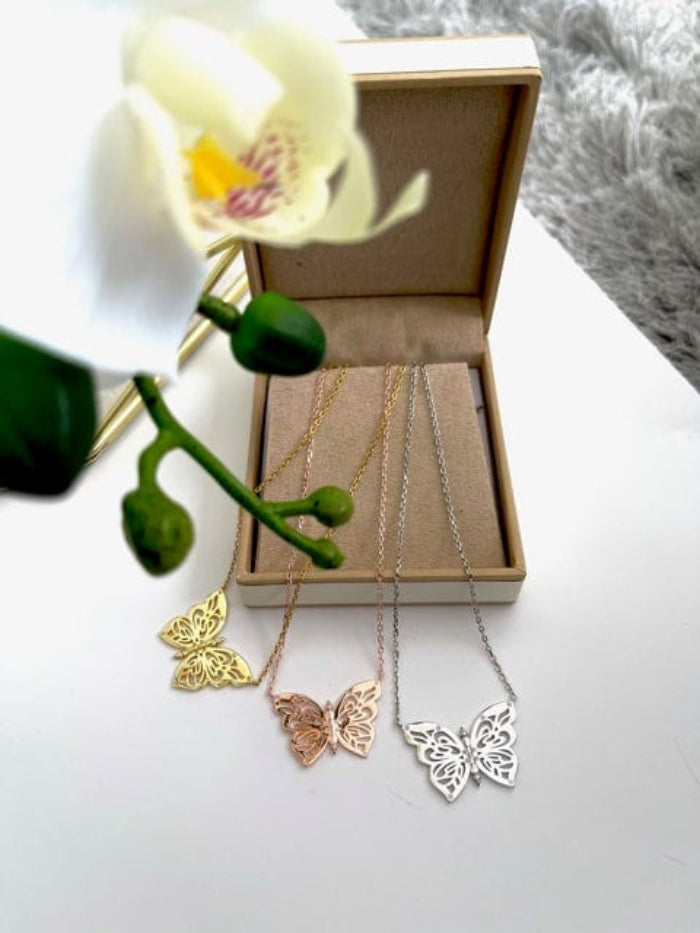 Butterfly-shaped necklaces in gold, rose gold, and silver on a white surface with a flower.