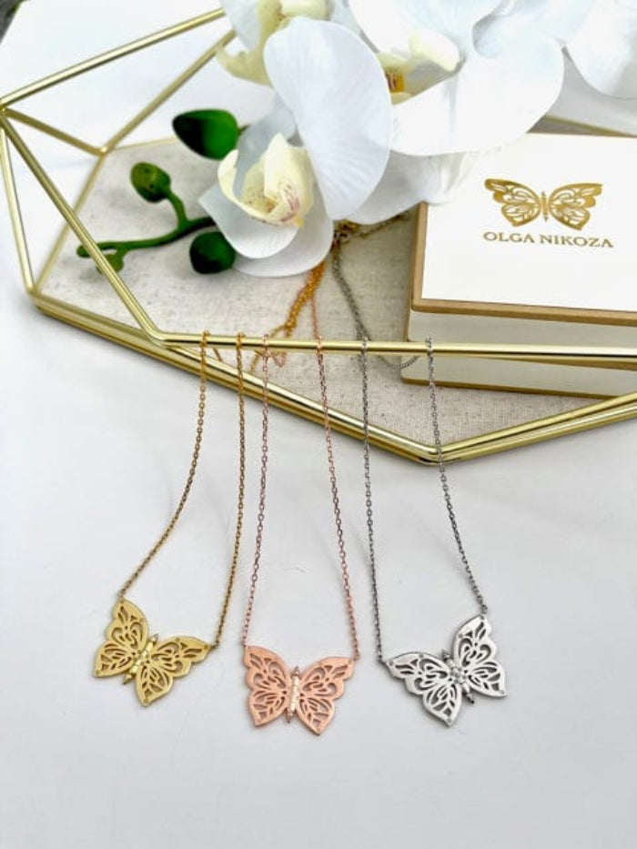 Three butterfly necklaces in gold, rose gold, and silver on a white surface with a branded box in the background.