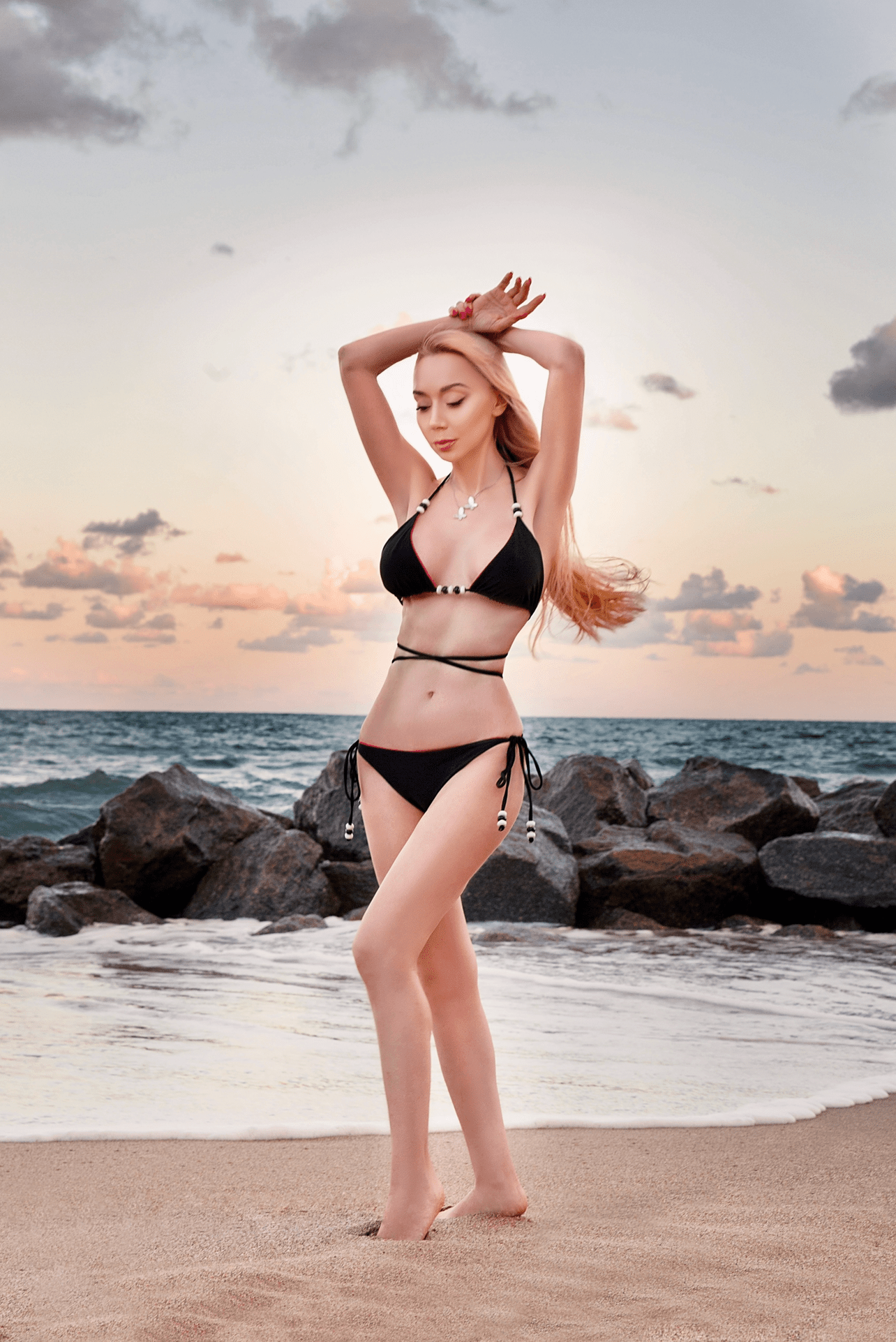 Woman in a black bikini standing on a beach with rocks and ocean in the background