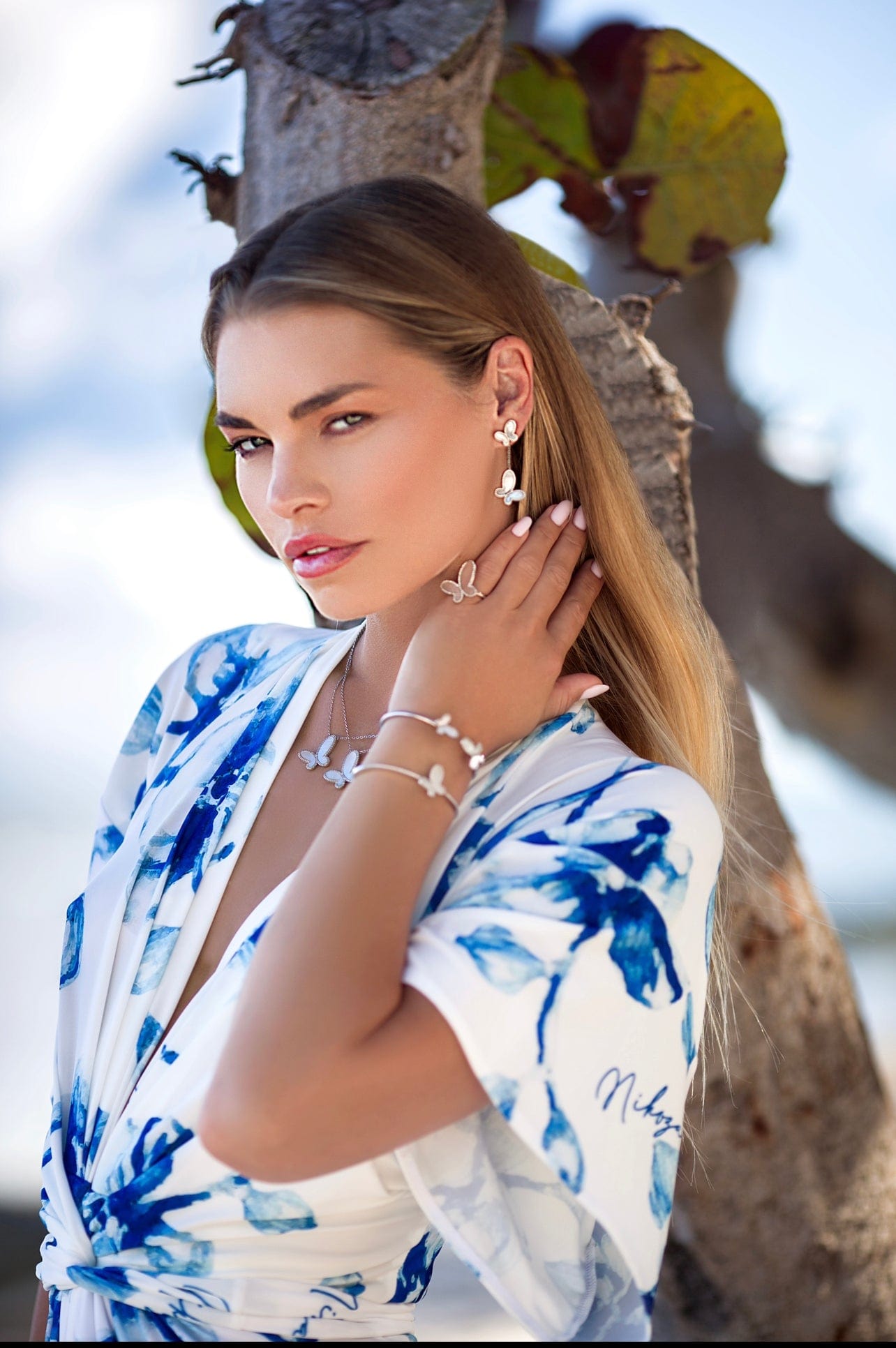 Woman wearing a blue and white floral dress with jewelry, standing against a natural background.