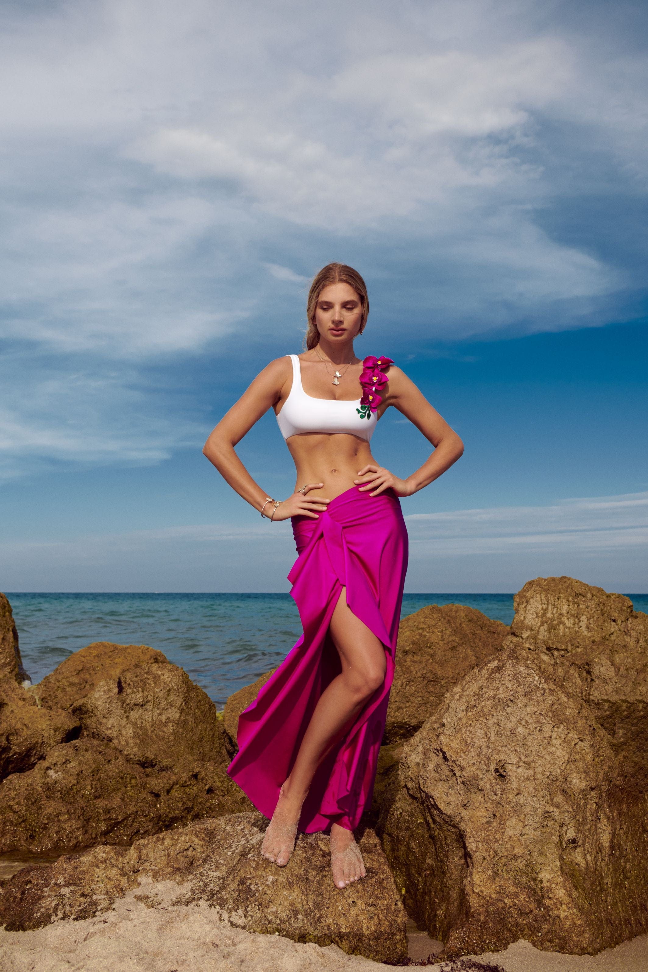 Woman in a white top and pink skirt standing on rocks by the ocean
