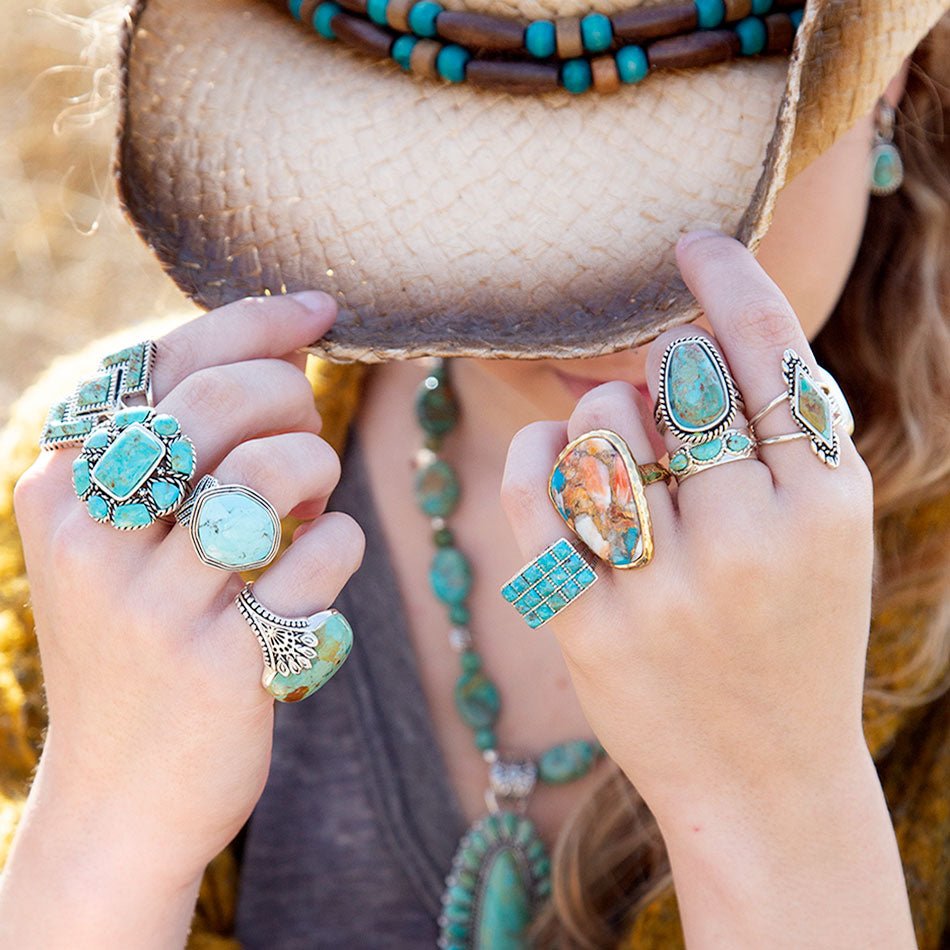 Person wearing multiple turquoise rings and a hat with beads, holding hands up.