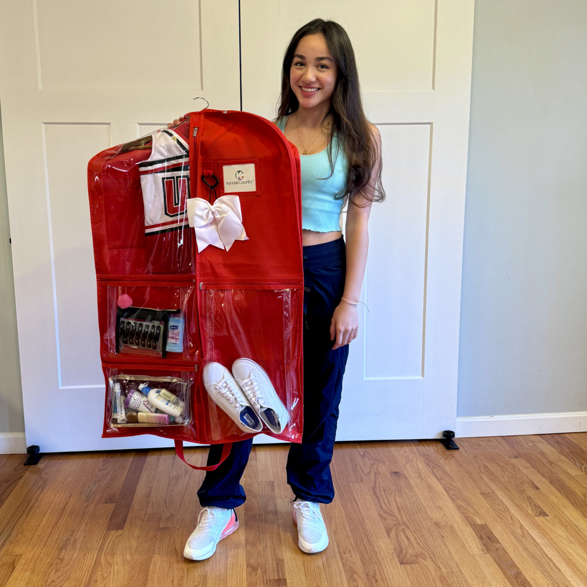 A young woman stands indoors holding a large red garment bag organizer filled with shoes, toiletries, and accessories. The room has light wood flooring and white paneled doors behind her.
