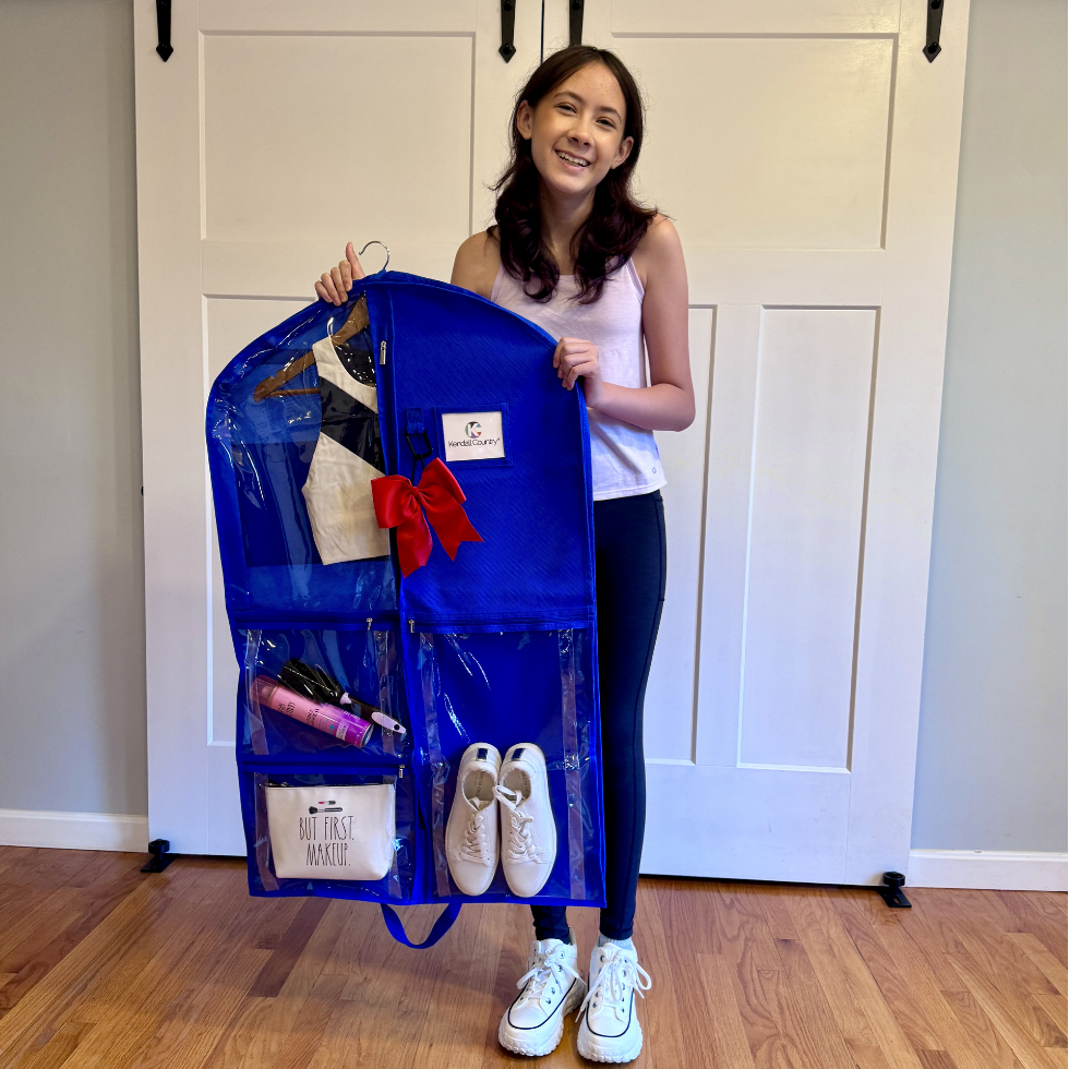 A woman stands in front of a white paneled door holding up a large blue hanging organizer bag filled with various items, including white sneakers, a small white bag, and a red bow. The organizer has multiple clear pockets and a white brand label that reads 'The Art of Magic'.